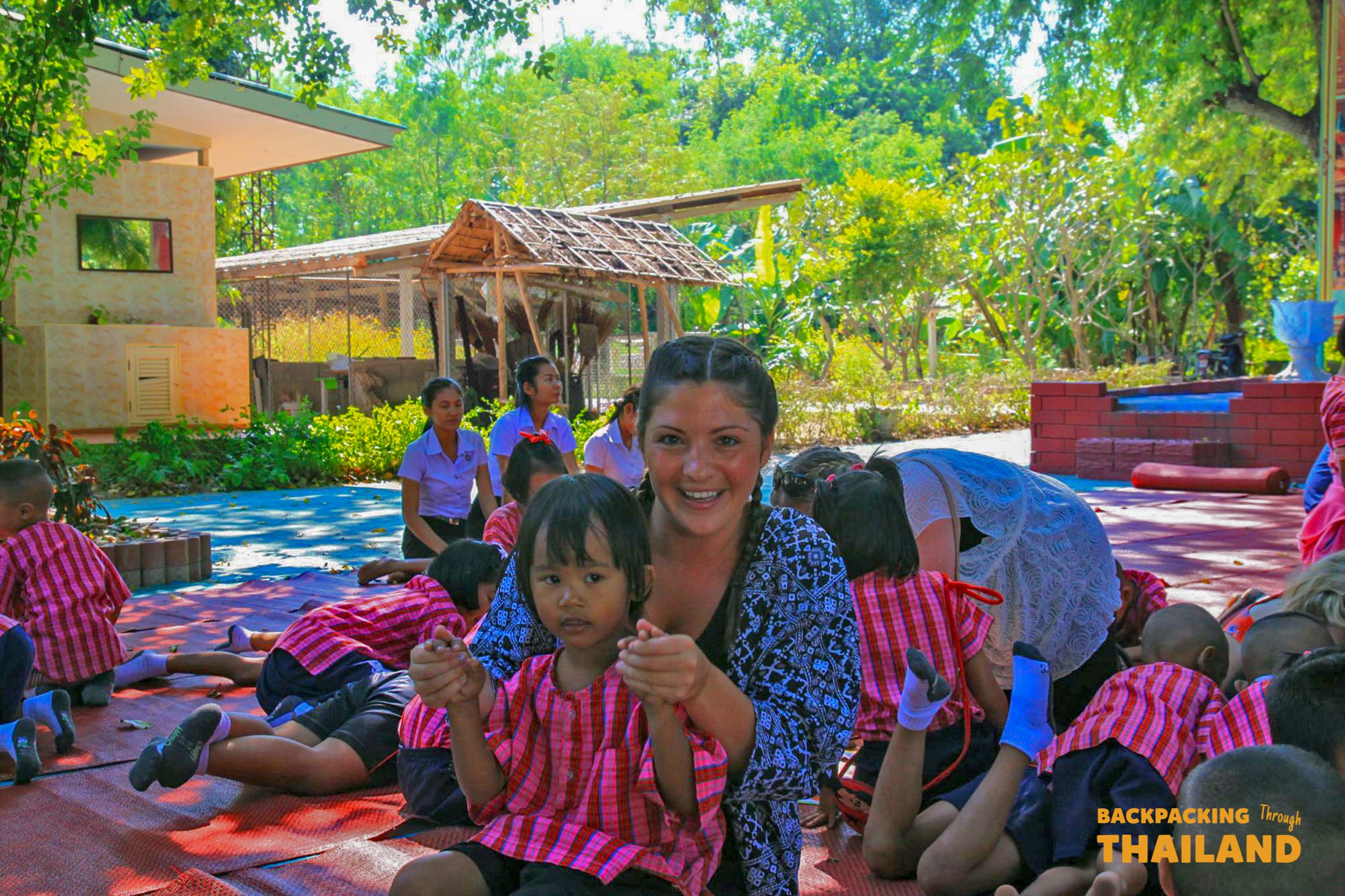 A backpacker smiles, holding hands with a young child during a visit to a local Thai school.