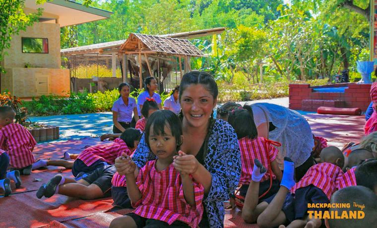 A backpacker smiles, holding hands with a young child during a visit to a local Thai school.