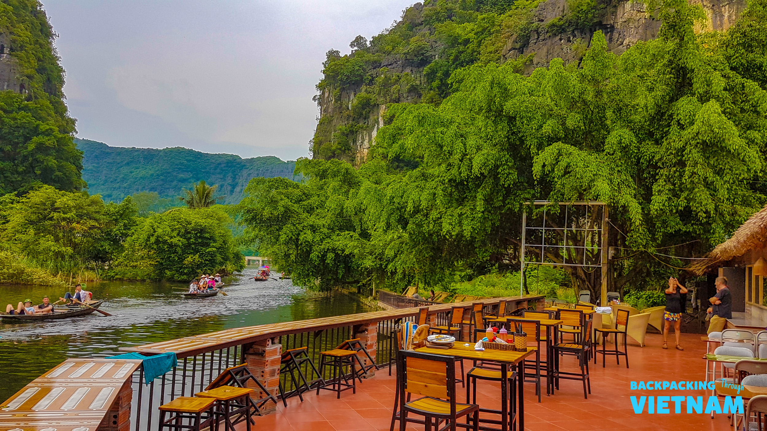 A scenic riverside restaurant patio in Tam Coc, with boats on the river and lush green mountains.