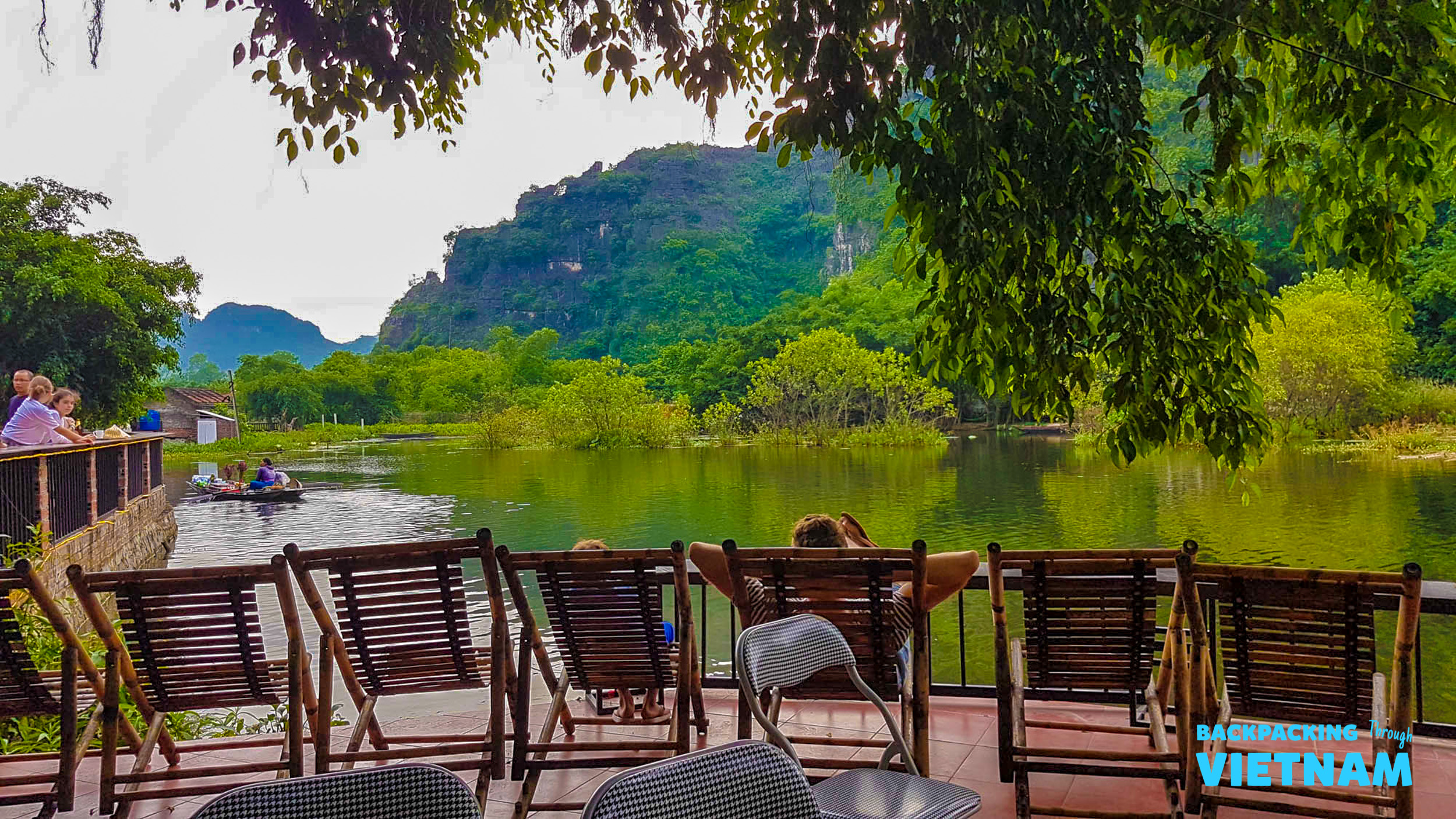 Travelers relax on a resort patio overlooking a serene lake with karstic mountains in Tam Coc, Vietnam.