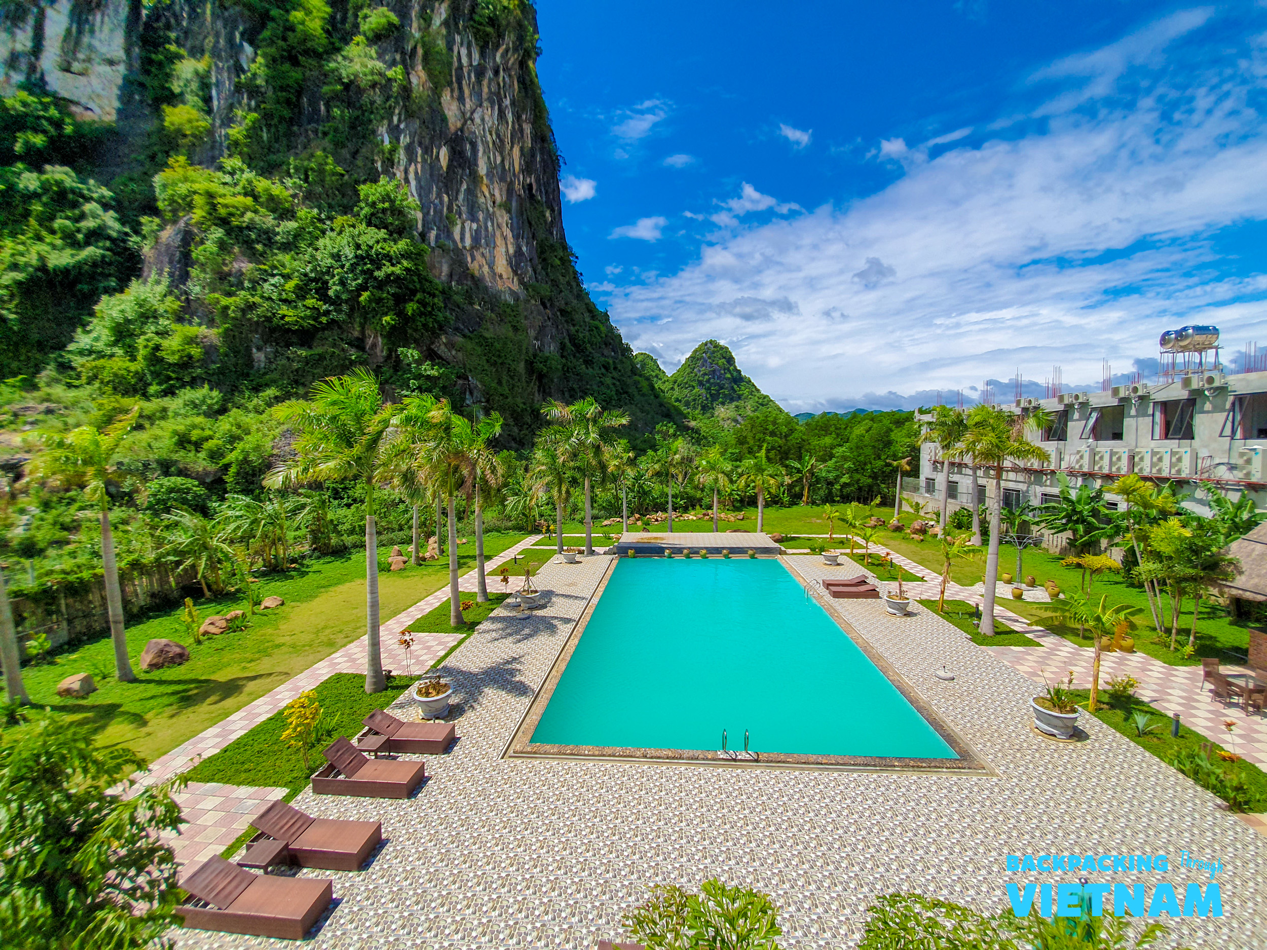 An inviting turquoise swimming pool with lounge chairs, palm trees, and karst mountains in Phong Nha.