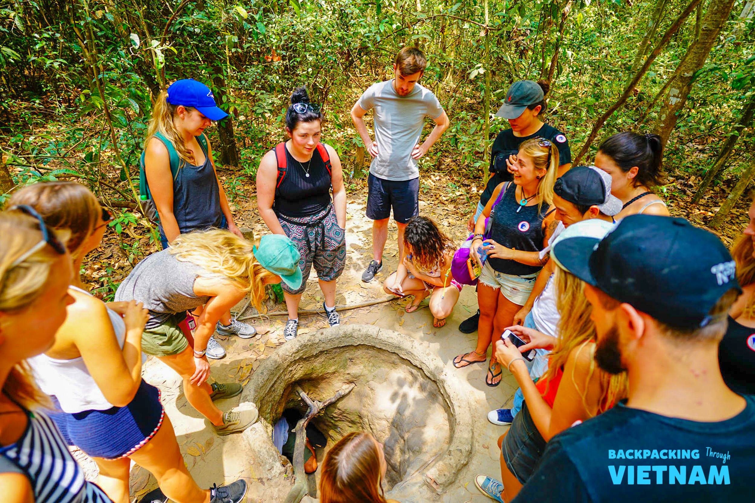 A group of backpackers gathers around a narrow tunnel entrance in a dense Vietnamese jungle.