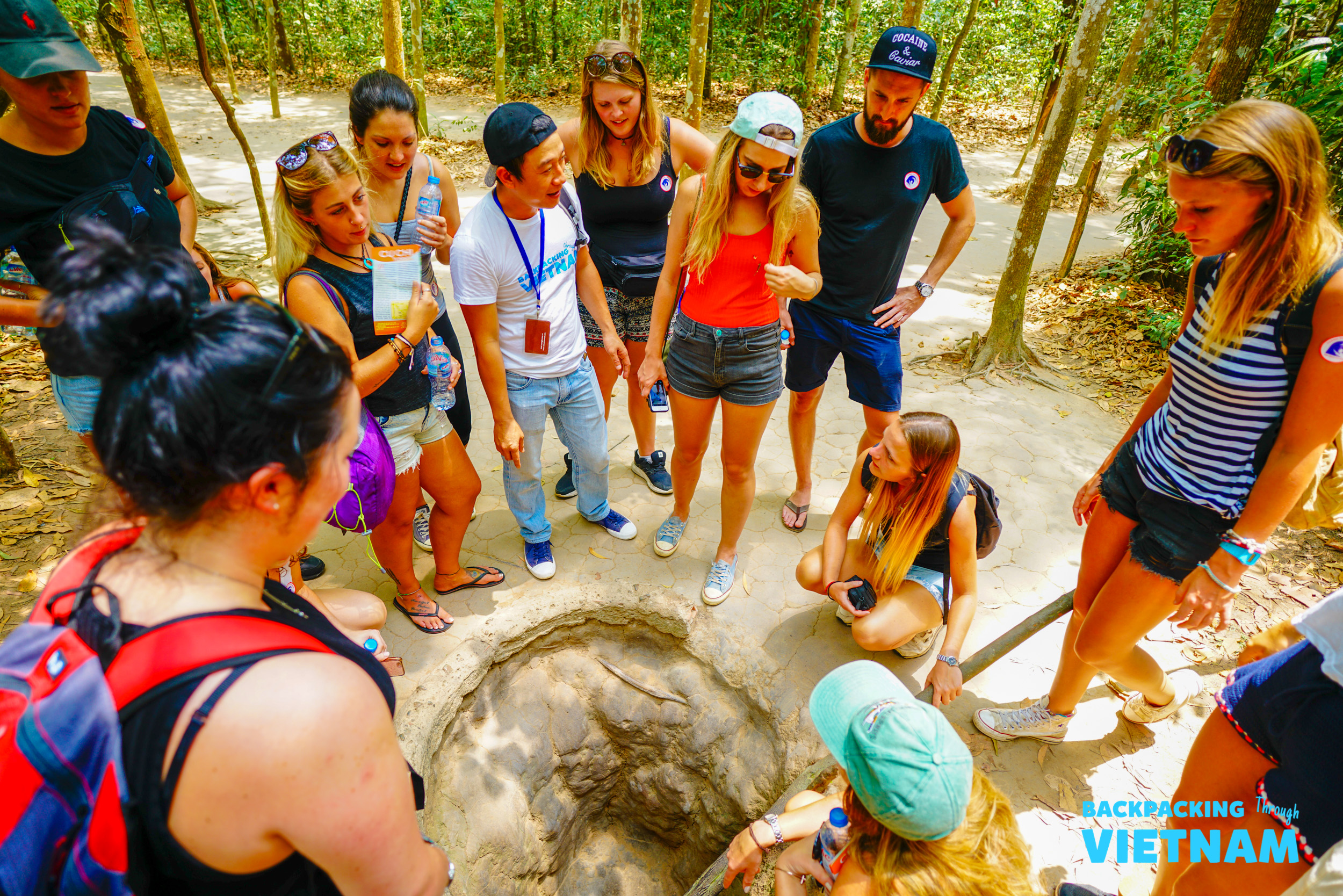 Tourists gather around a narrow tunnel entrance, learning about the Cu Chi Tunnels in Vietnam.
