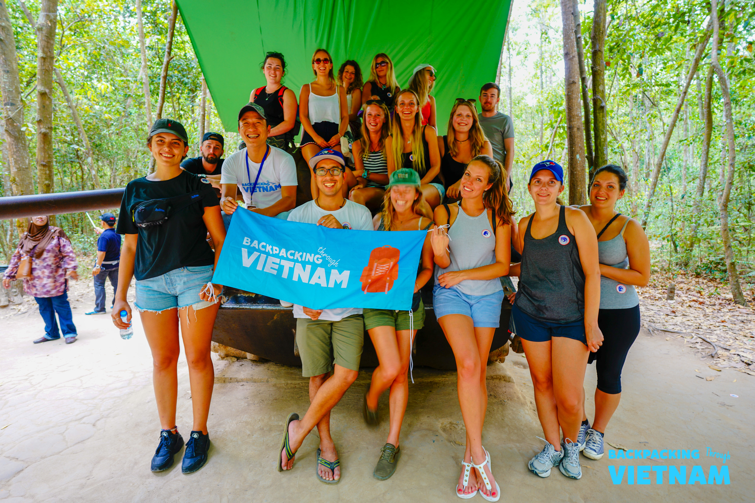 A large group of happy backpackers posing with their Backpacking Vietnam flag in a jungle.