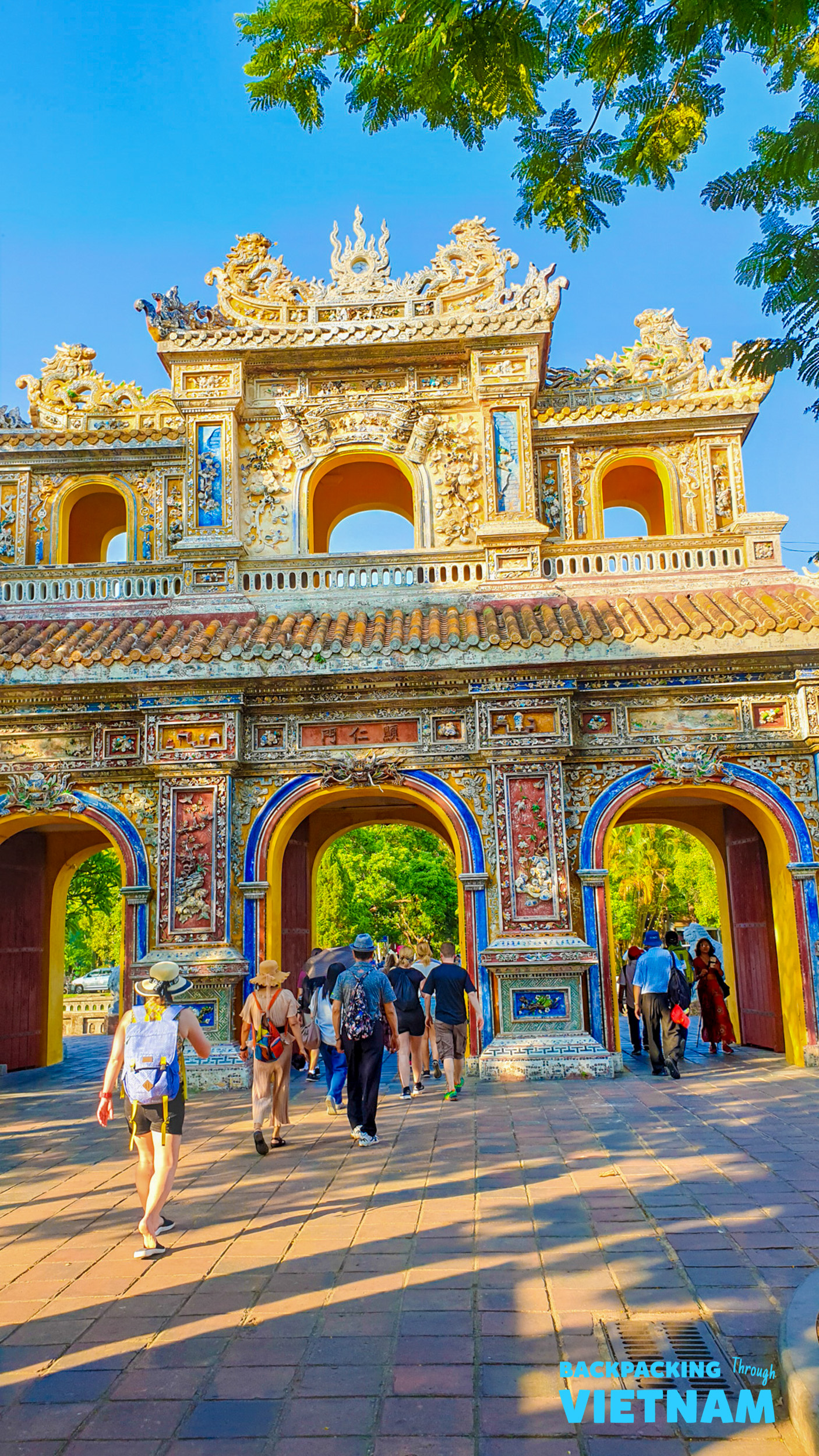 Backpackers explore the grand, colorful Imperial City gate in Hue, Vietnam under a clear blue sky.