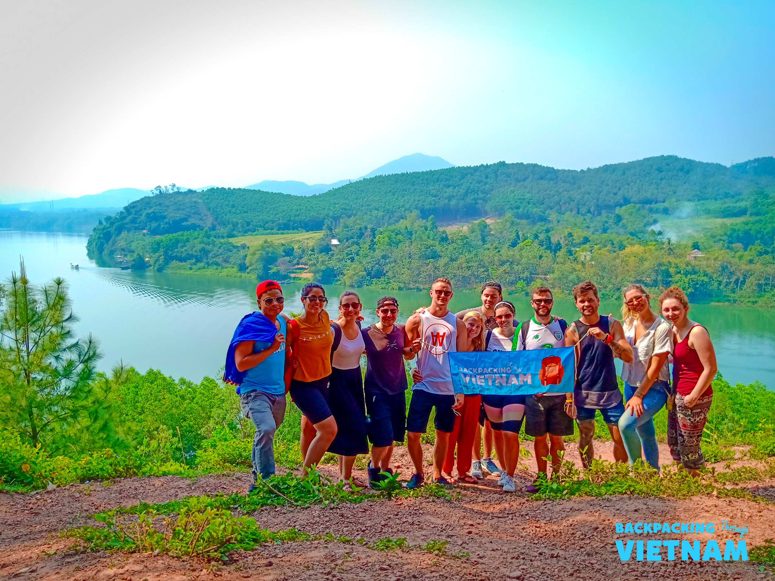 Happy travelers gather for a group photo by a serene lake surrounded by lush green hills in Hue.
