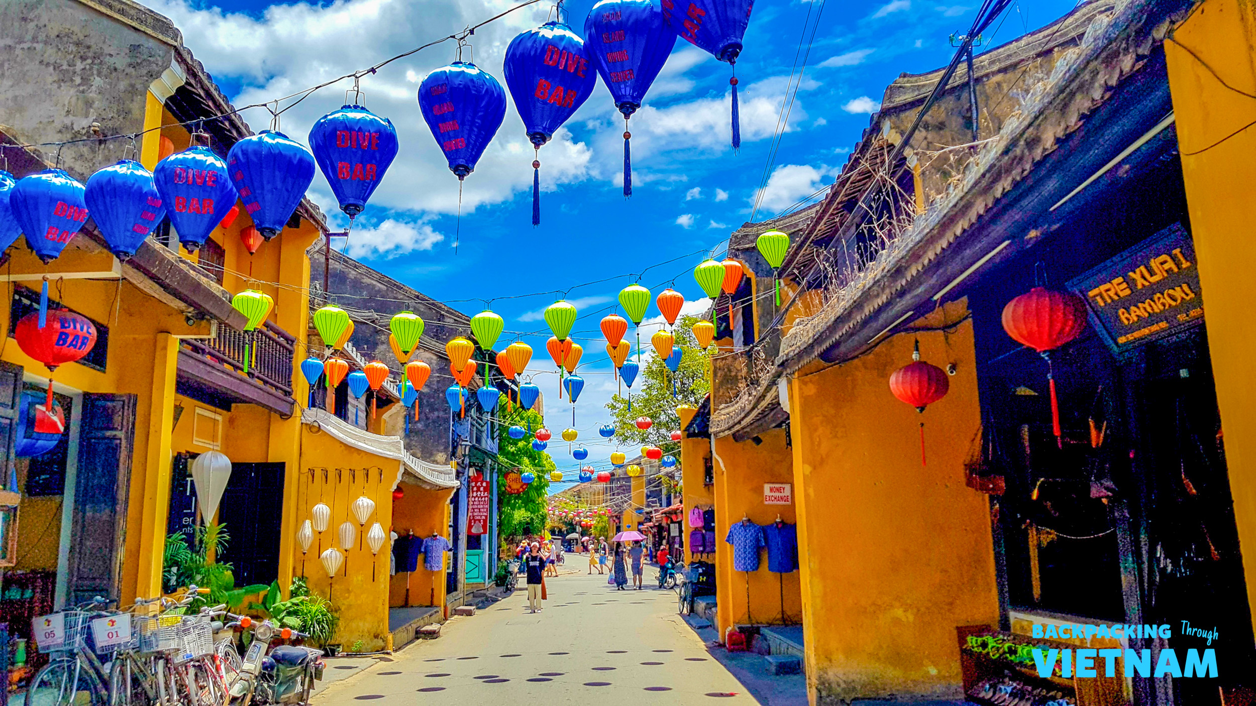 Colorful lanterns illuminate a bustling street in Hoi An, Vietnam, under a bright blue sky.