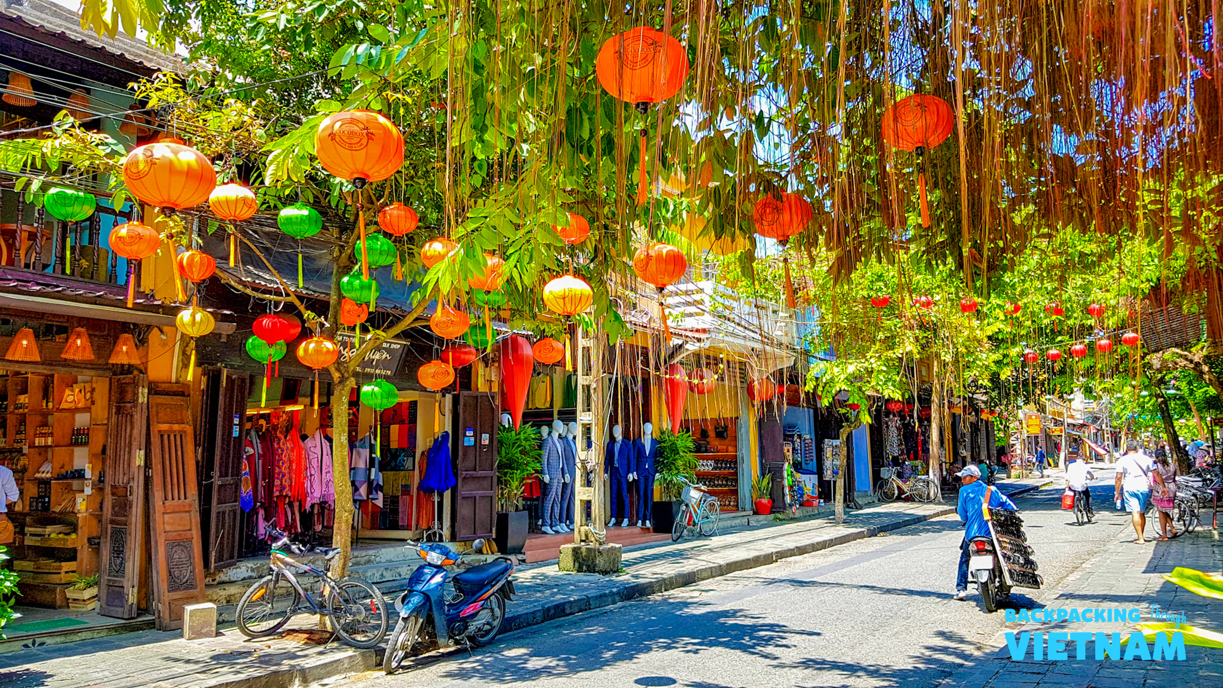 Colorful lanterns hang over a bustling street in Hoi An, Vietnam, with shops and people.