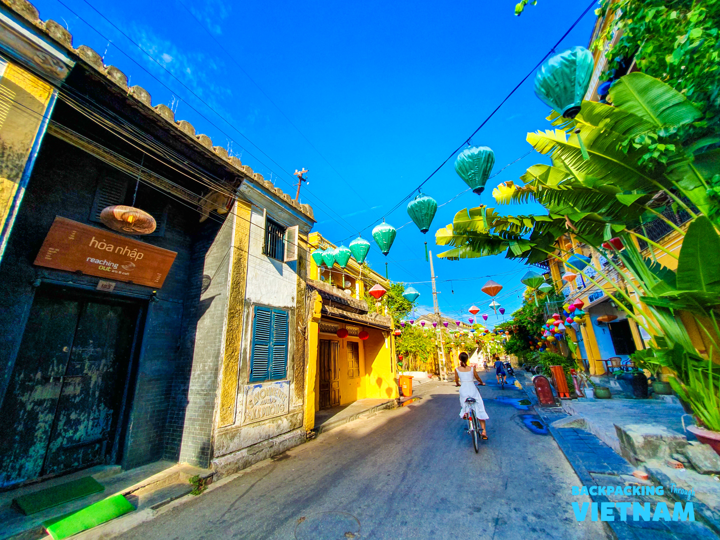 A person cycles down a vibrant street in Hoi An, Vietnam, adorned with colorful lanterns.