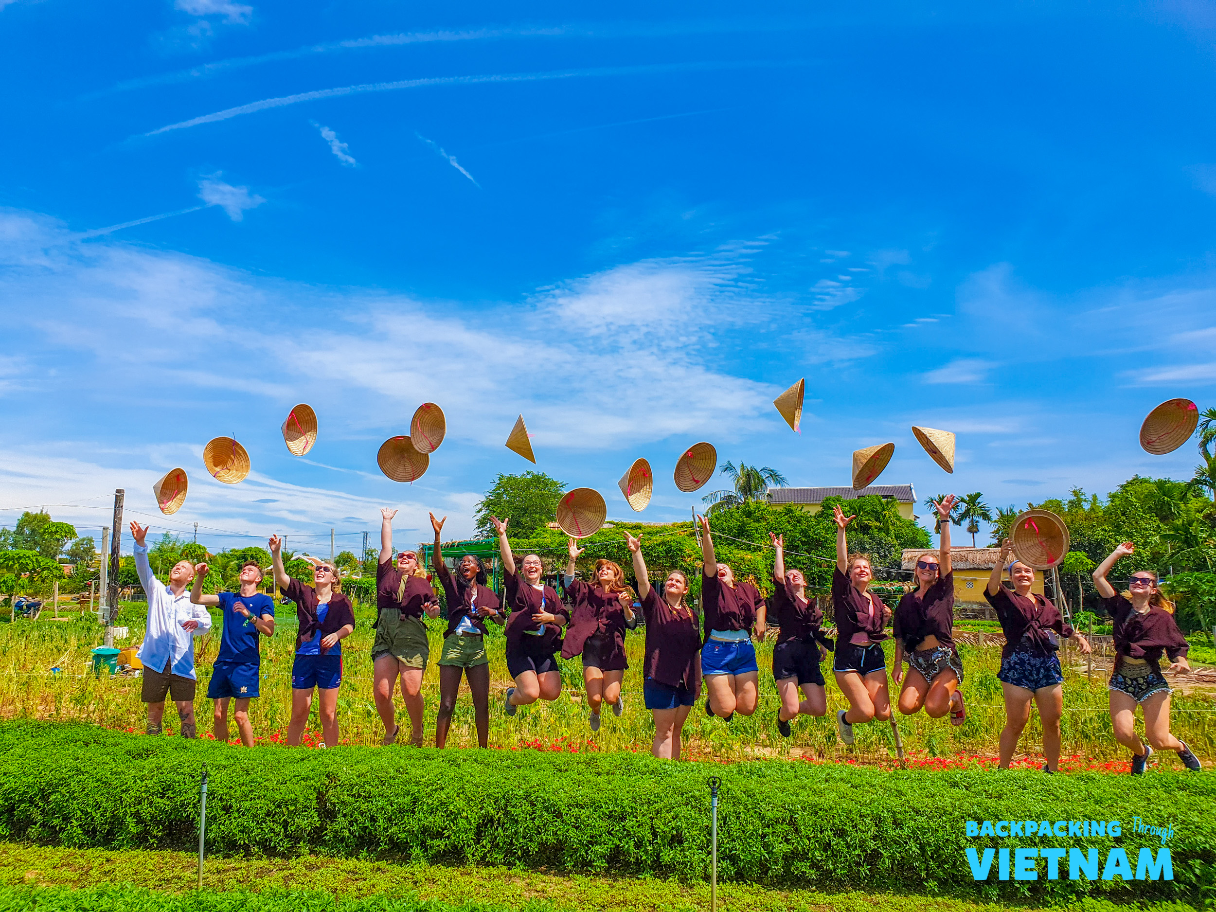 Joyful travelers jump, tossing traditional conical hats in a vibrant green field under a blue sky, Hoi An.