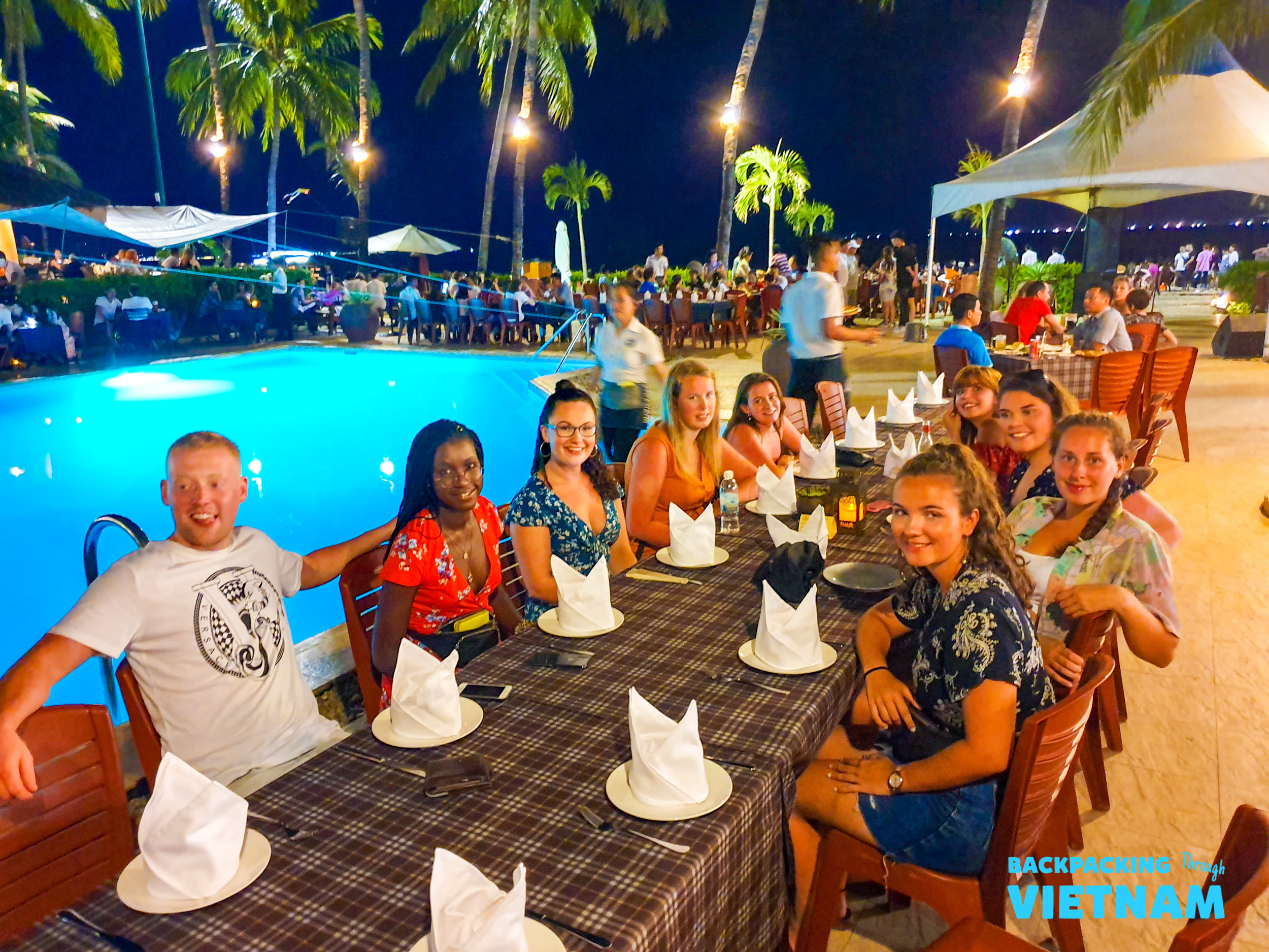 A group of happy backpackers enjoying a lively outdoor dinner by a pool in Nha Trang.