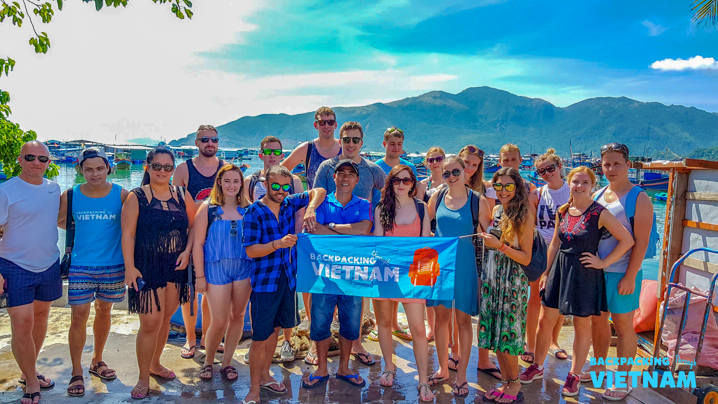 Smiling backpackers hold a "Backpacking Vietnam" banner by the sea under a bright sky in Nha Trang.