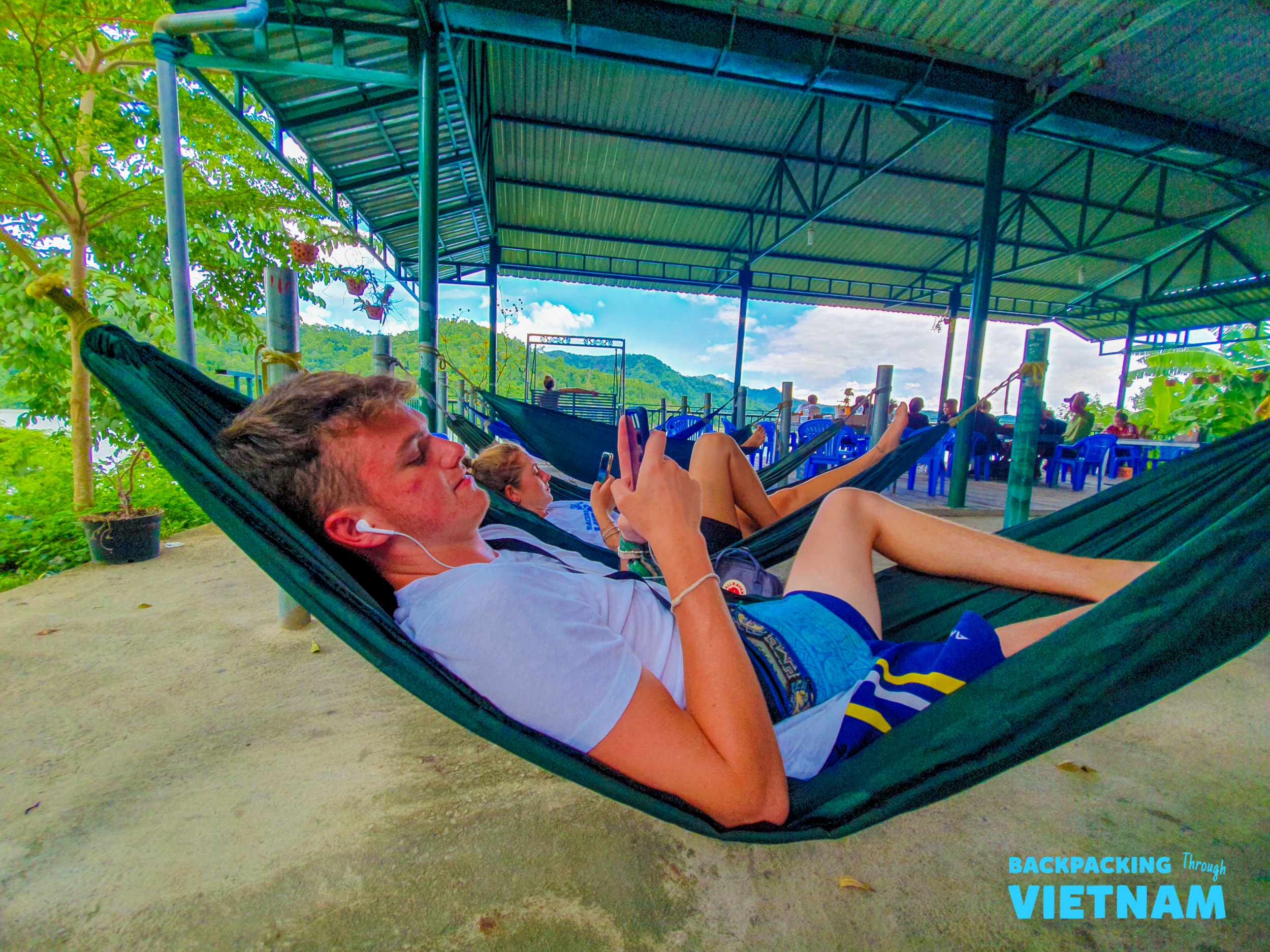A young man relaxes in a green hammock, using his phone with earphones in scenic Dalat, Vietnam.