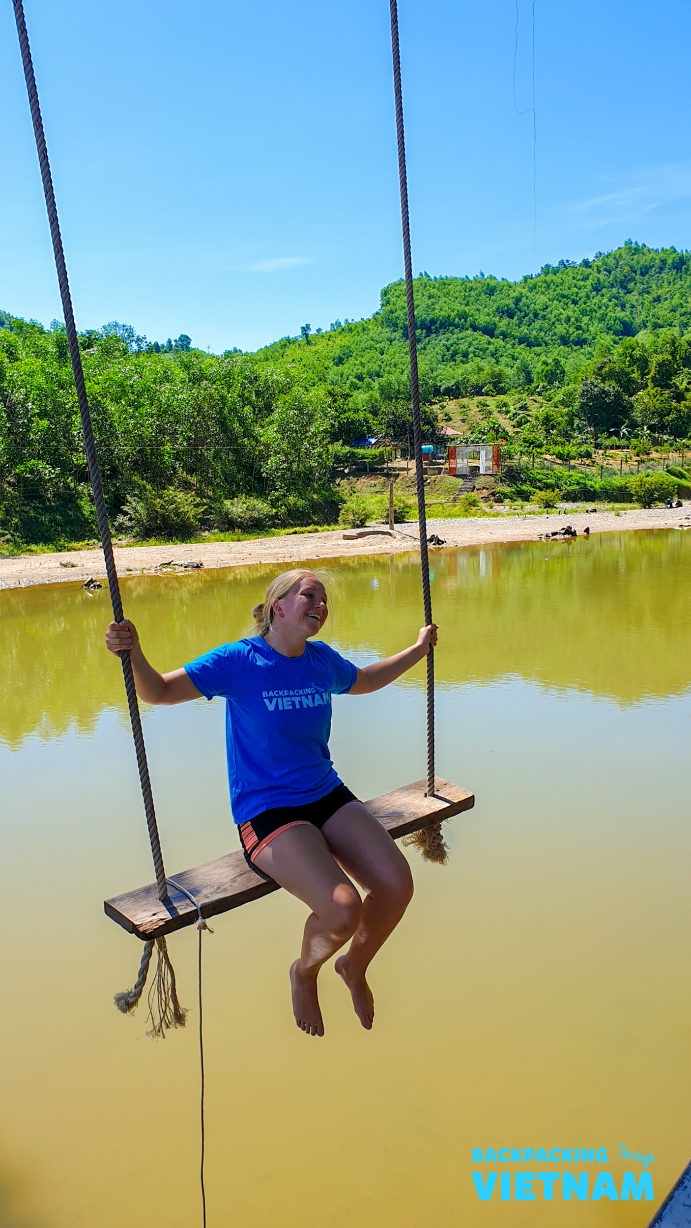 A happy backpacker swings over a lake in Dalat, surrounded by lush green hills under a clear sky.