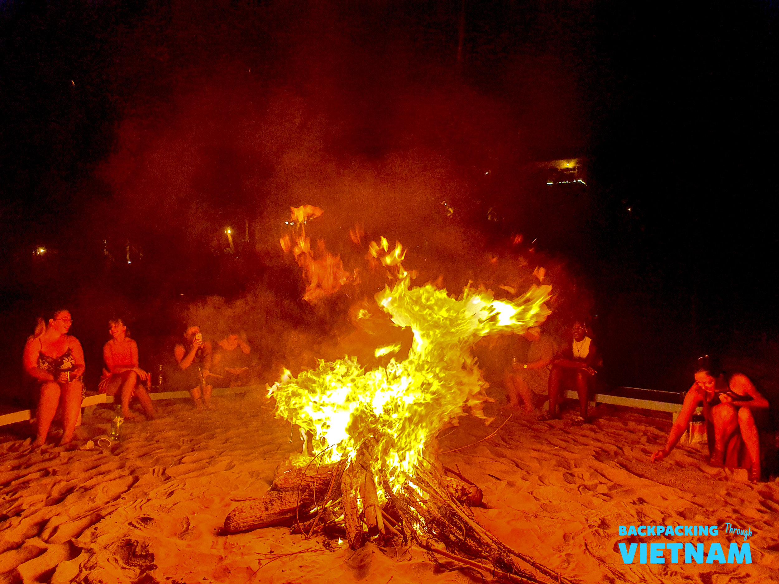 Backpackers gather around a large, crackling bonfire on a sandy beach at night in Mui Ne.