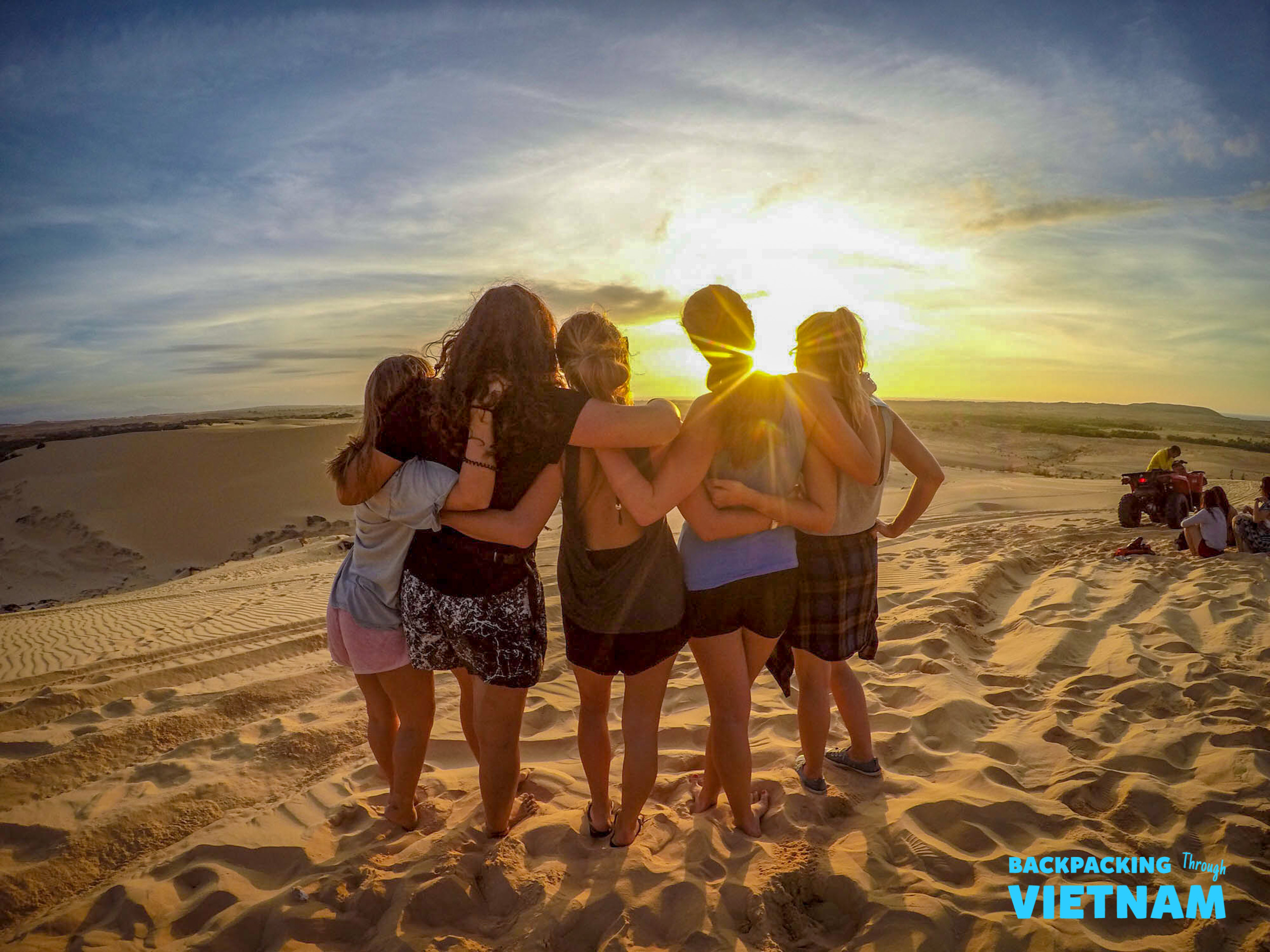 Five friends embrace on sand dunes, watching a beautiful golden sunrise in Mui Ne, Vietnam.
