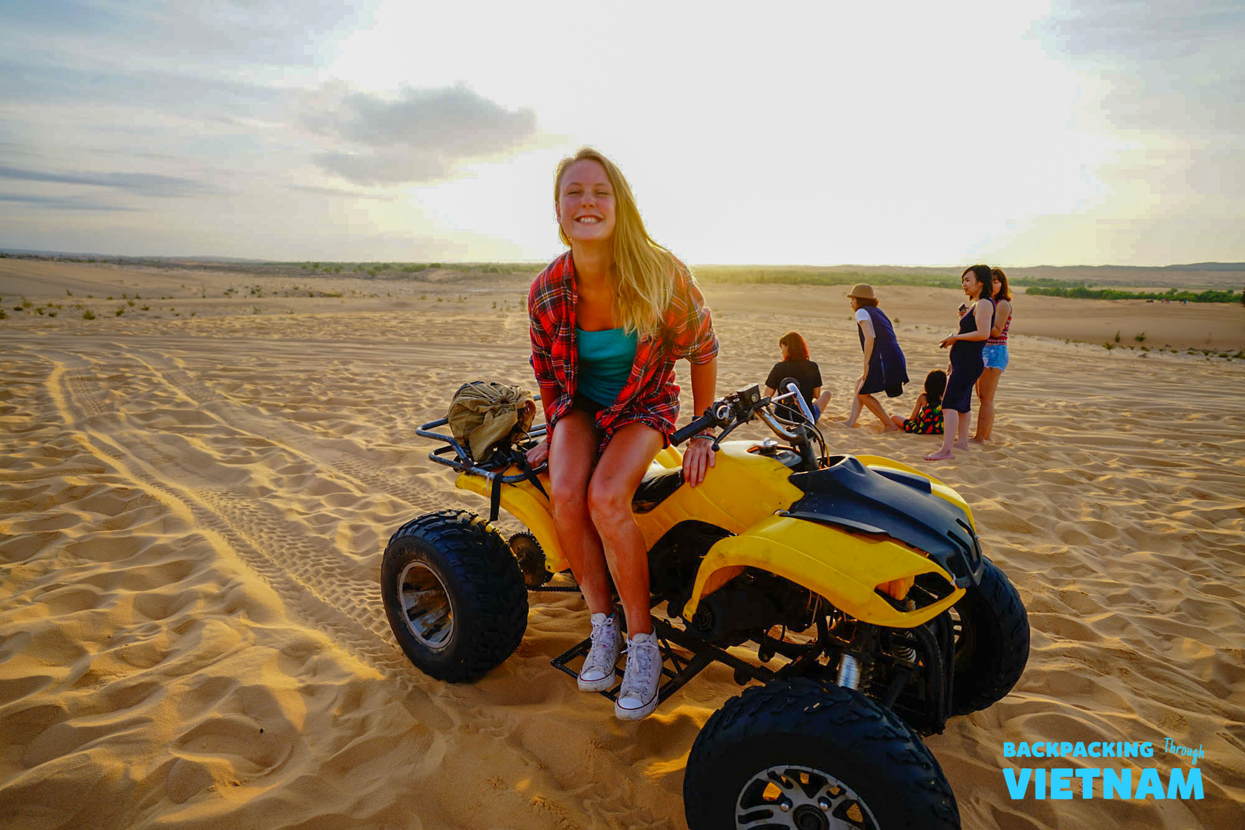 A smiling woman sits on a yellow ATV on vast sand dunes at sunrise in Mui Ne, Vietnam.