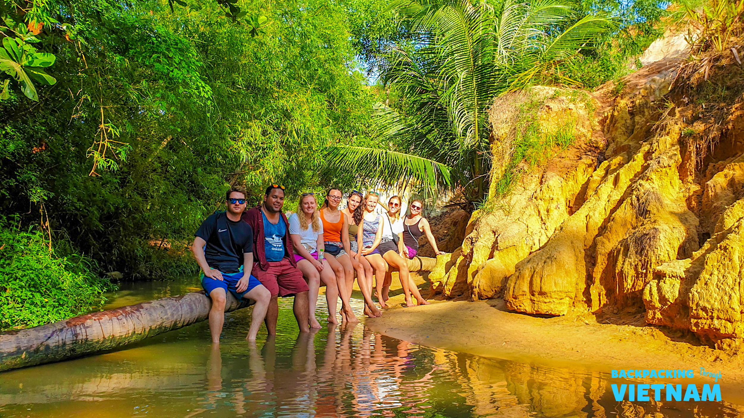 Eight happy backpackers sit on a fallen log over the Fairy Stream in Mui Ne, Vietnam.