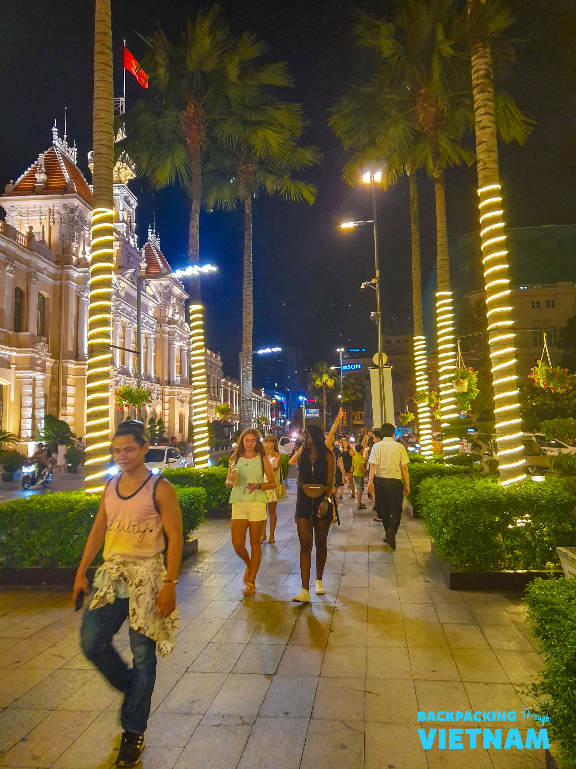 Friends enjoy a lively evening stroll past illuminated palm trees and historic buildings in Ho Chi Minh.