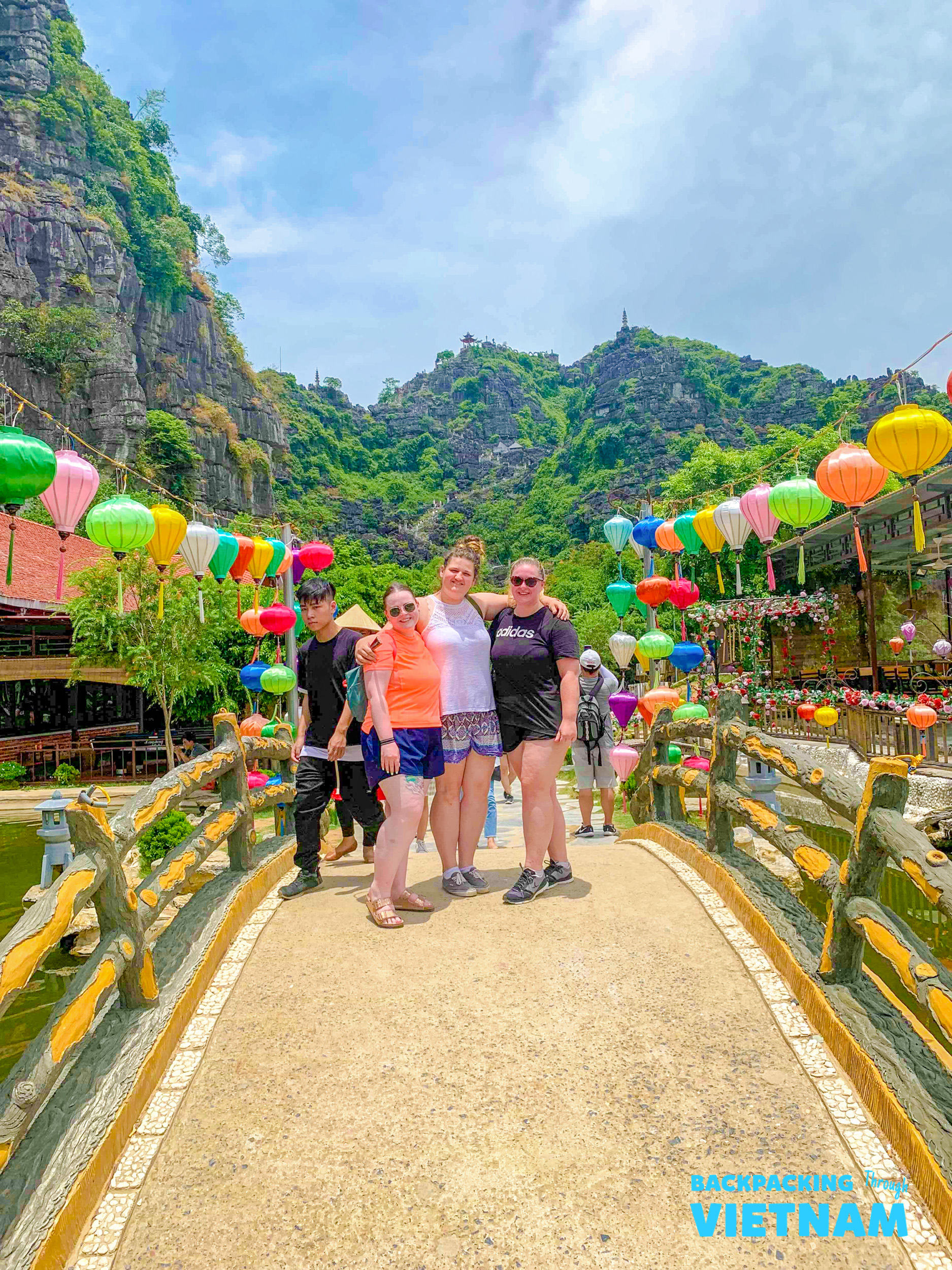 Four happy backpackers pose on a colorful bridge with limestone mountains and lanterns in Tam Coc, Vietnam.