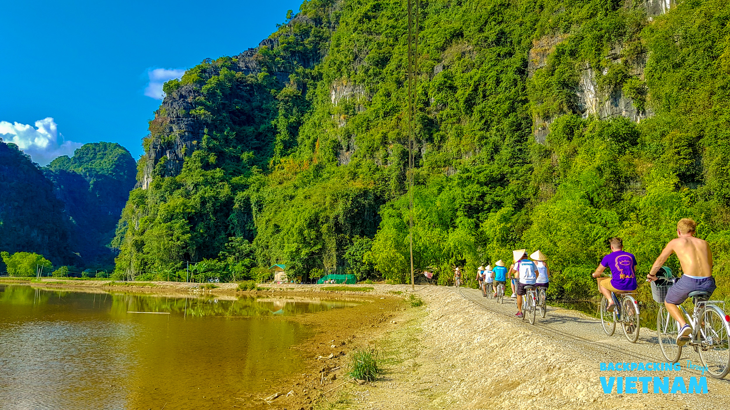 Tourists cycle along a scenic path beside a river with towering green mountains in Tam Coc, Vietnam.
