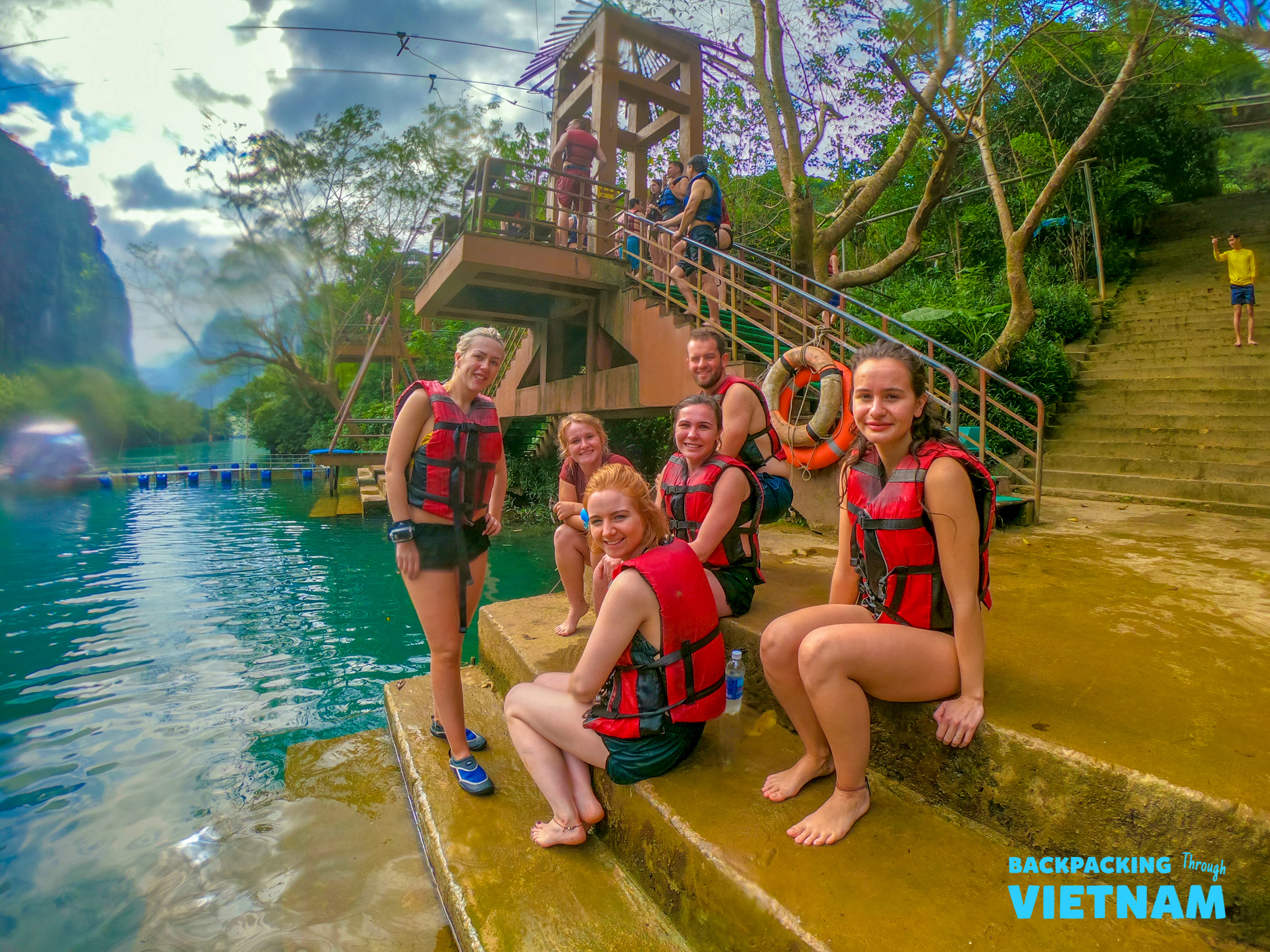 Happy backpackers in life vests on steps beside clear blue water, ready for adventure in Phong Nha.