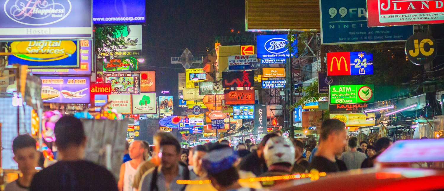 People stroll through a bustling Bangkok street illuminated by countless vibrant neon signs at night.