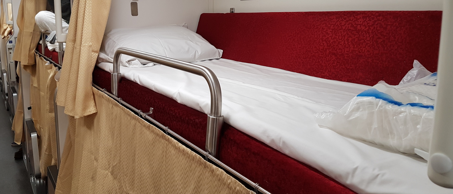 A clean, comfortable bunk bed with a red mattress and white sheets on a Thai sleeper train.