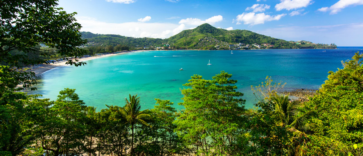 Panoramic view of Patong Beach in Phuket, Thailand, with clear turquoise water and lush hills.
