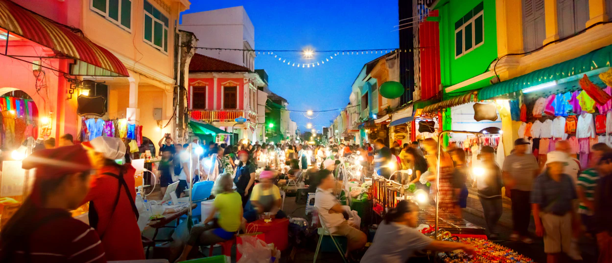 A lively night market in Phuket Old Town, with crowds of people browsing colorful stalls and bright lights.
