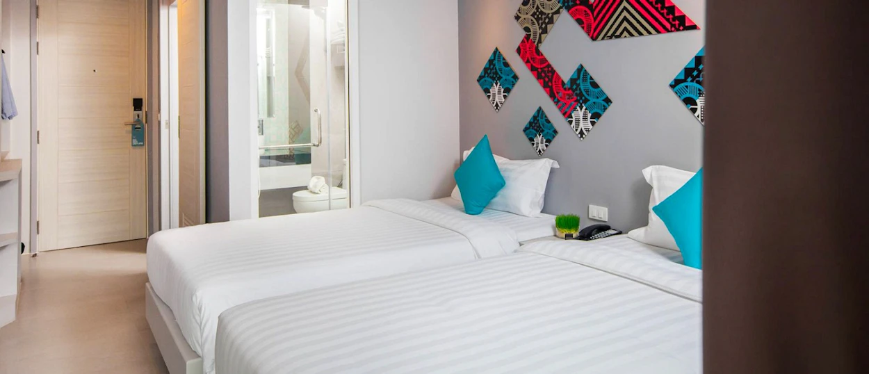 A bright, modern hotel room in Phuket with two single beds, blue pillows, and colorful wall art.
