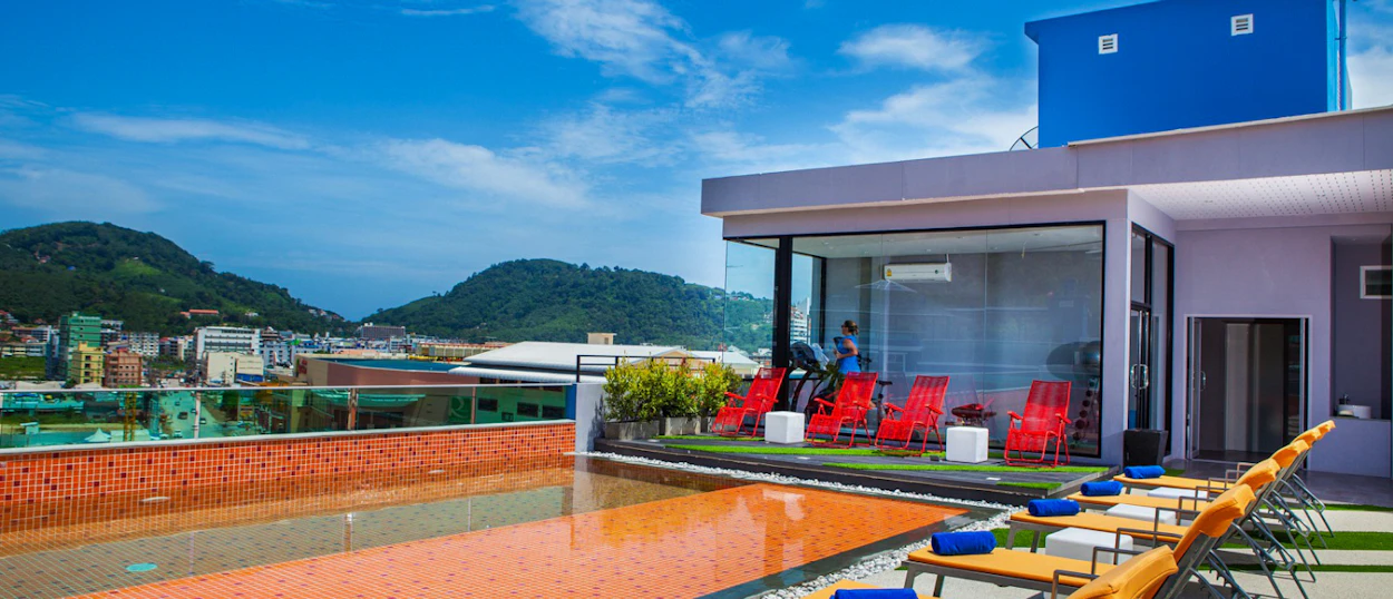 Sunny rooftop swimming pool with orange lounge chairs overlooking green hills and city buildings in Phuket.