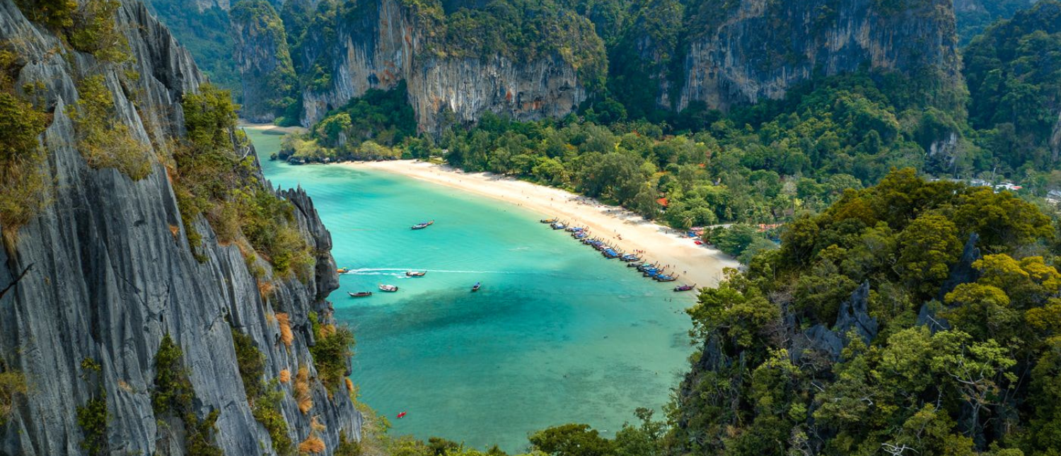 Overhead view of Railay Beach with turquoise water, white sand, longtail boats, and towering limestone cliffs.