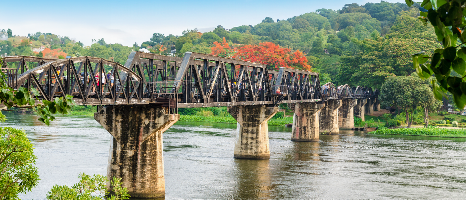 People walk across the historic River Kwai Bridge over a wide river in Kanchanaburi, Thailand.