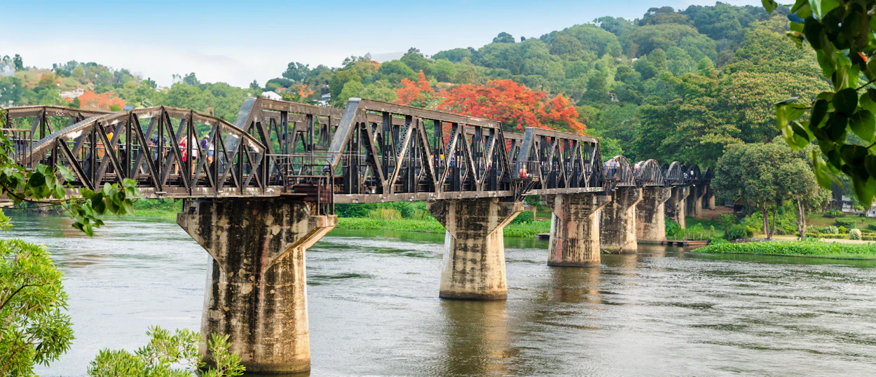 People walk across the historic River Kwai Bridge over a wide river in Kanchanaburi, Thailand.