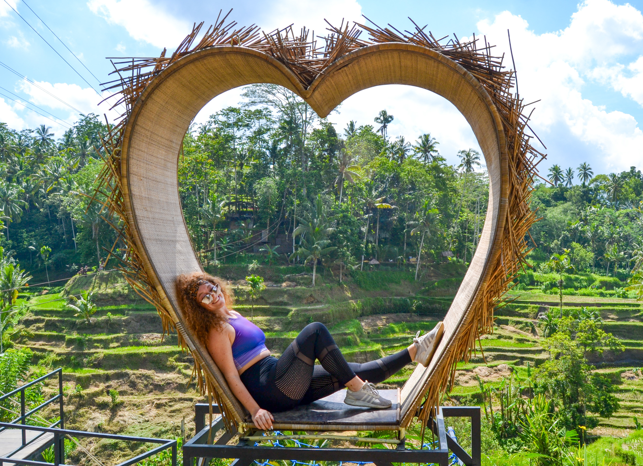 A smiling woman relaxing inside a heart-shaped bamboo structure overlooking Bali's green rice terraces.