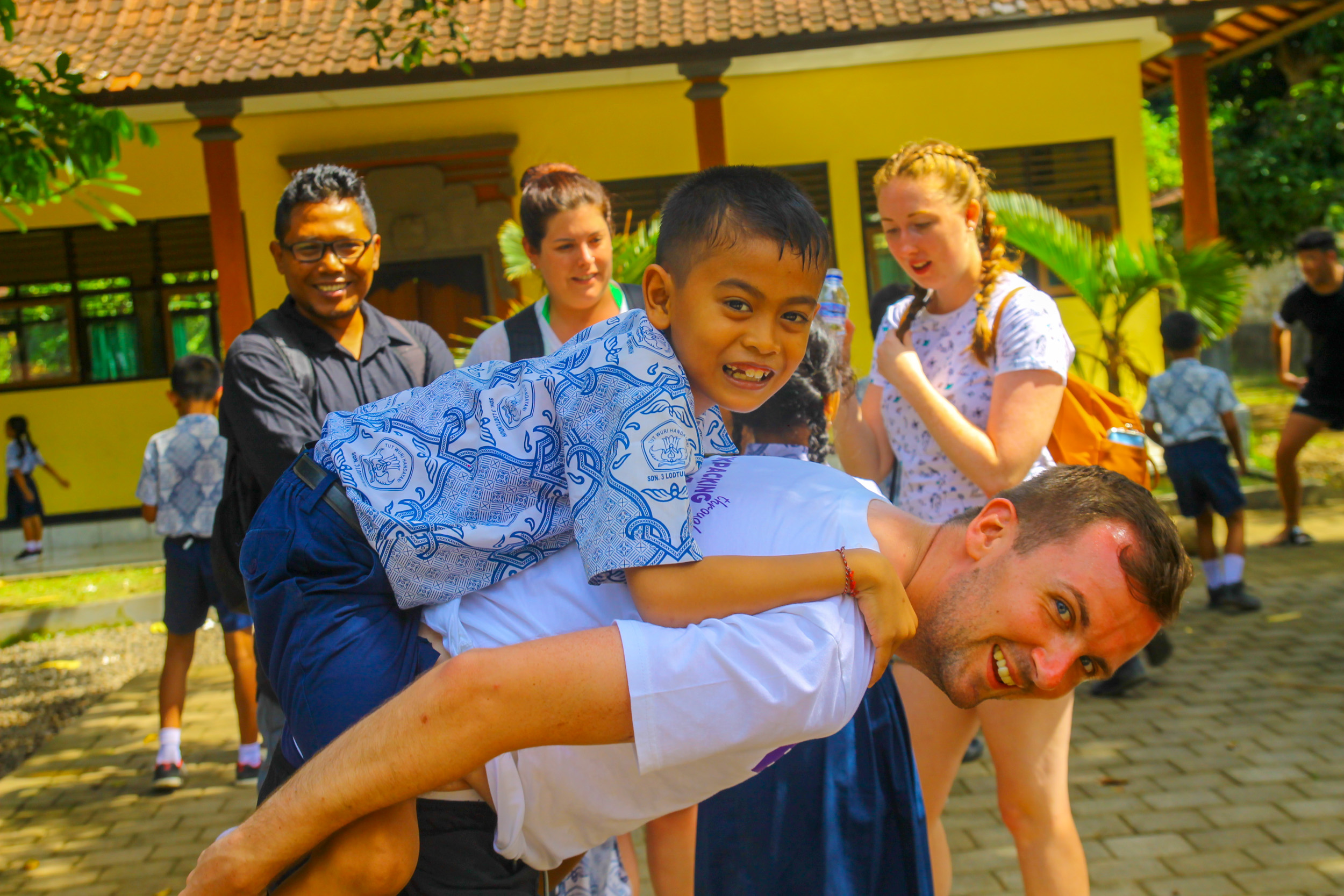 A joyful man gives a piggyback ride to a smiling boy in a sunny Bali village.