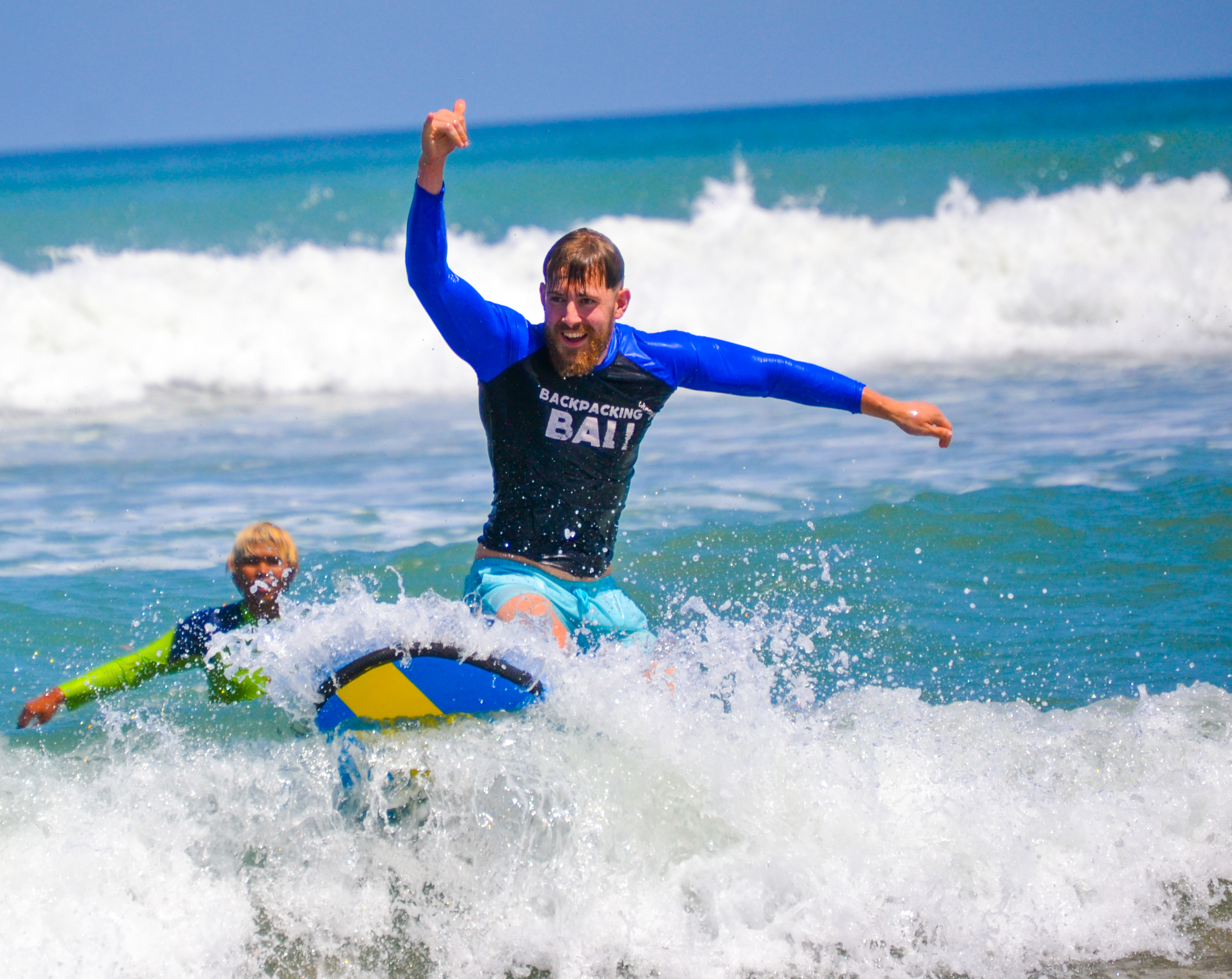 Joyful man in a 'Backpacking Bali' shirt rides a skimboard on a wave, thumbs-up.