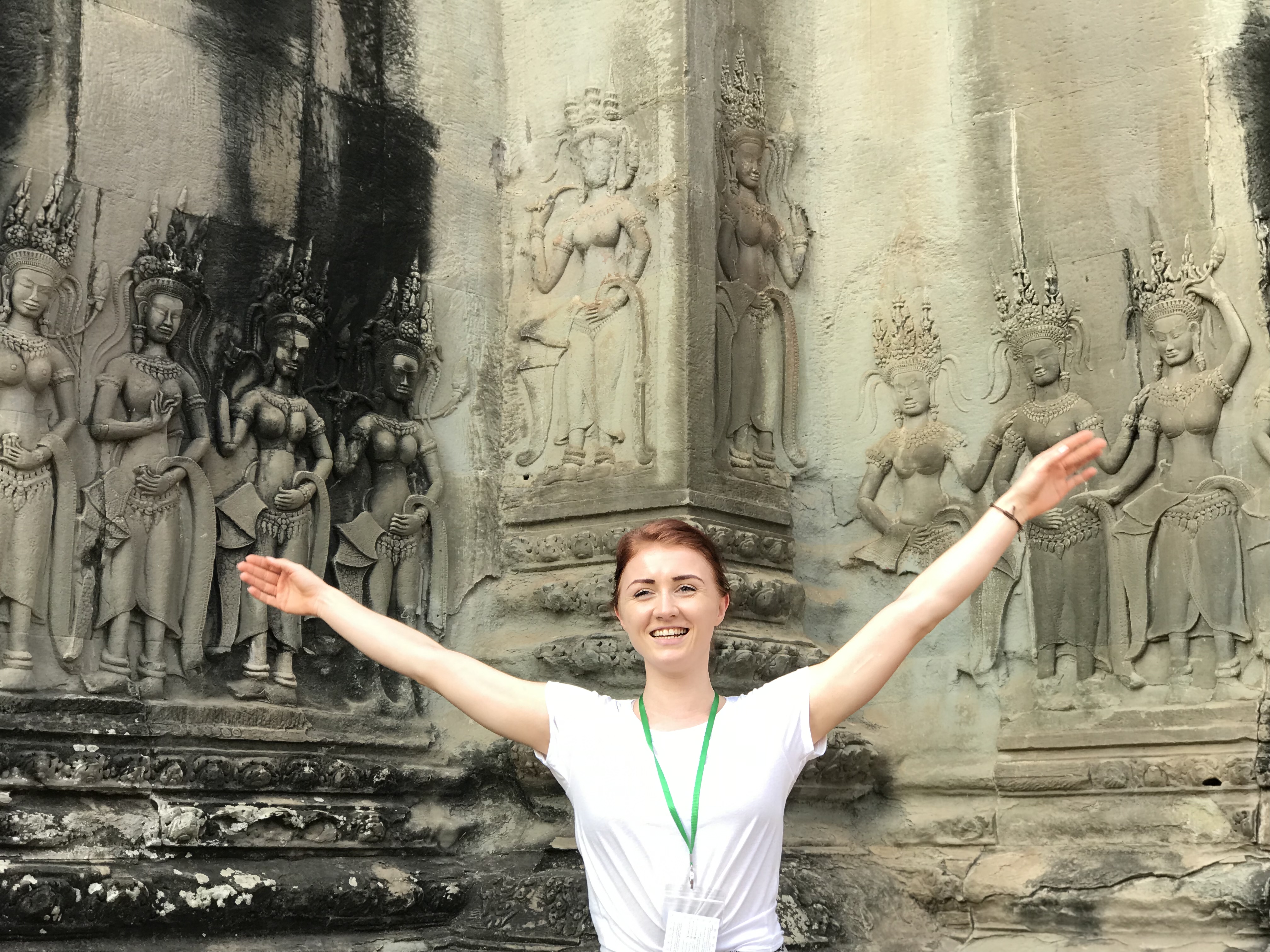 Smiling woman with outstretched arms in front of ancient Angkor Wat temple carvings.