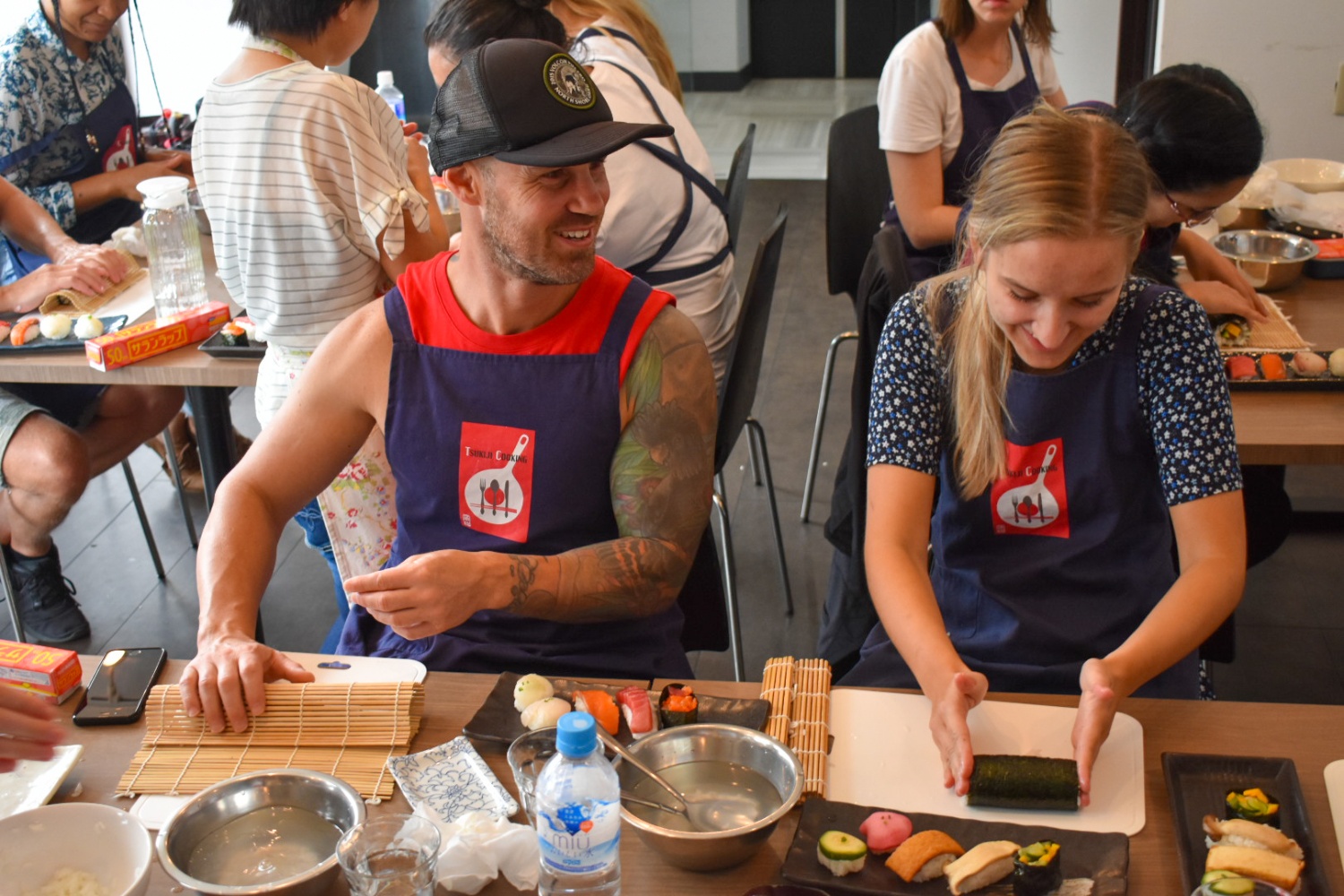 Two happy travelers in aprons learning to make sushi rolls at a cooking class in Japan.