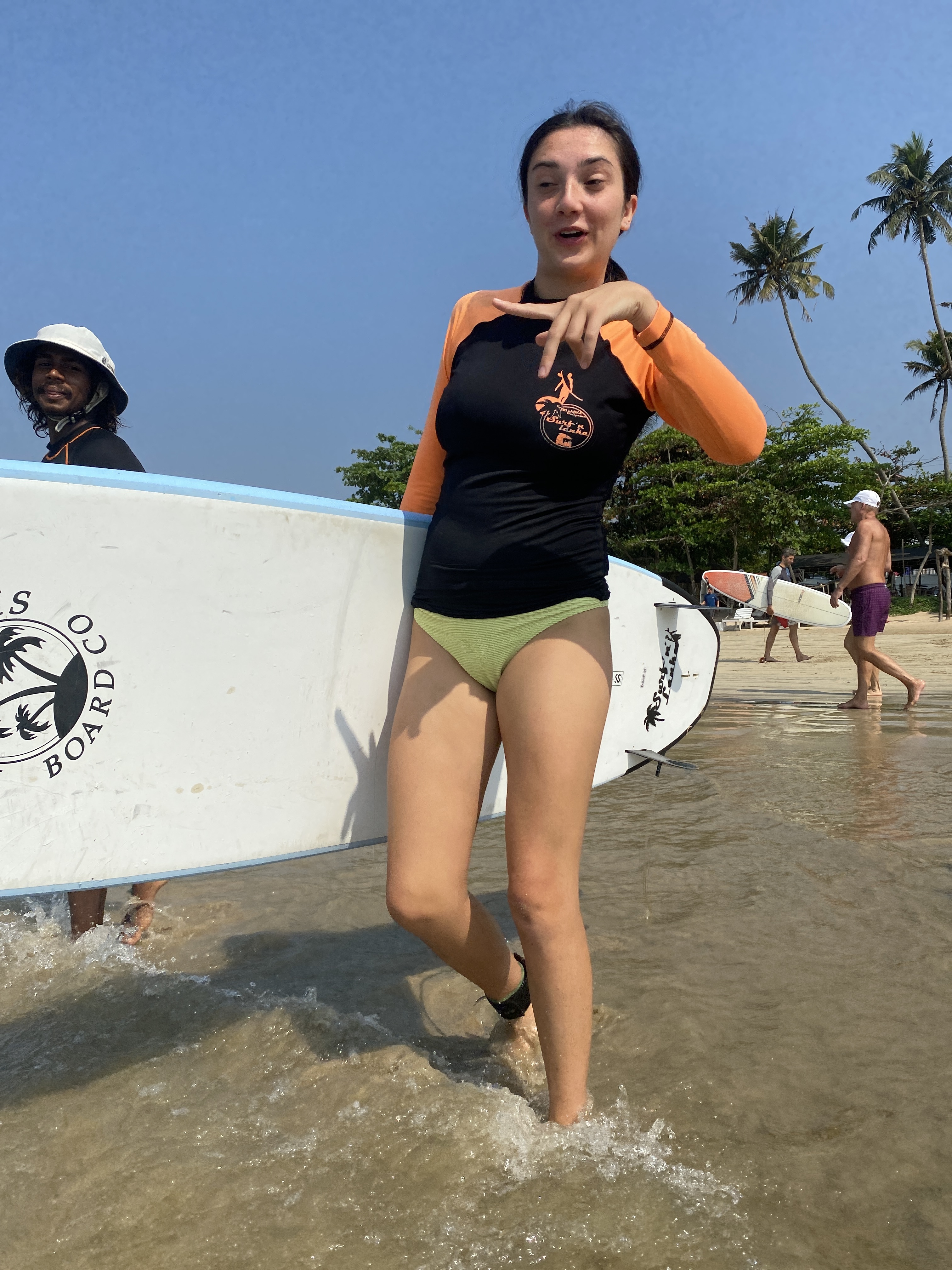 Smiling woman in a rash guard walks from the ocean with a surfboard on a sunny Sri Lankan beach.