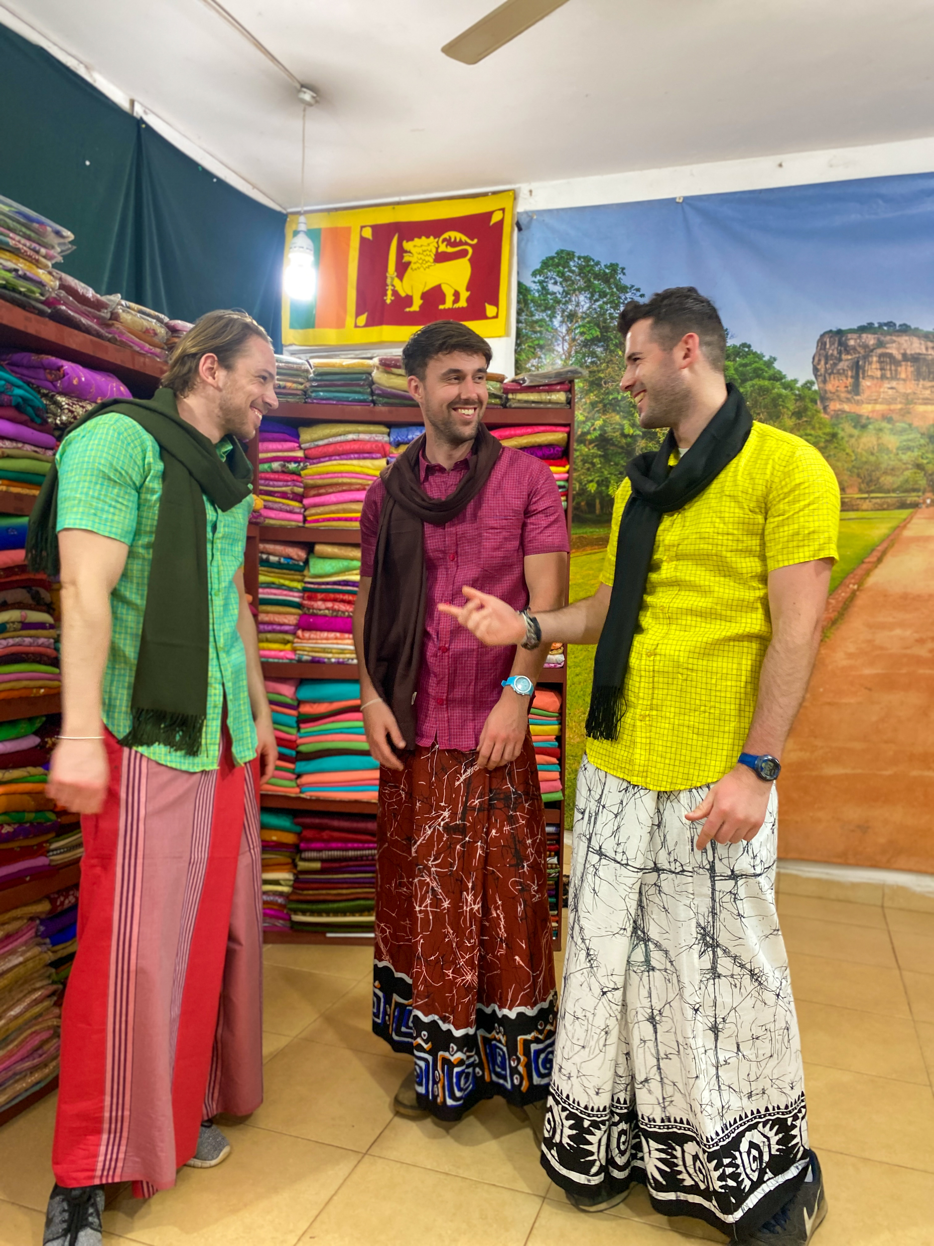 Three friends laughing, trying on colorful traditional sarongs and scarves in a vibrant Sri Lankan textile shop.