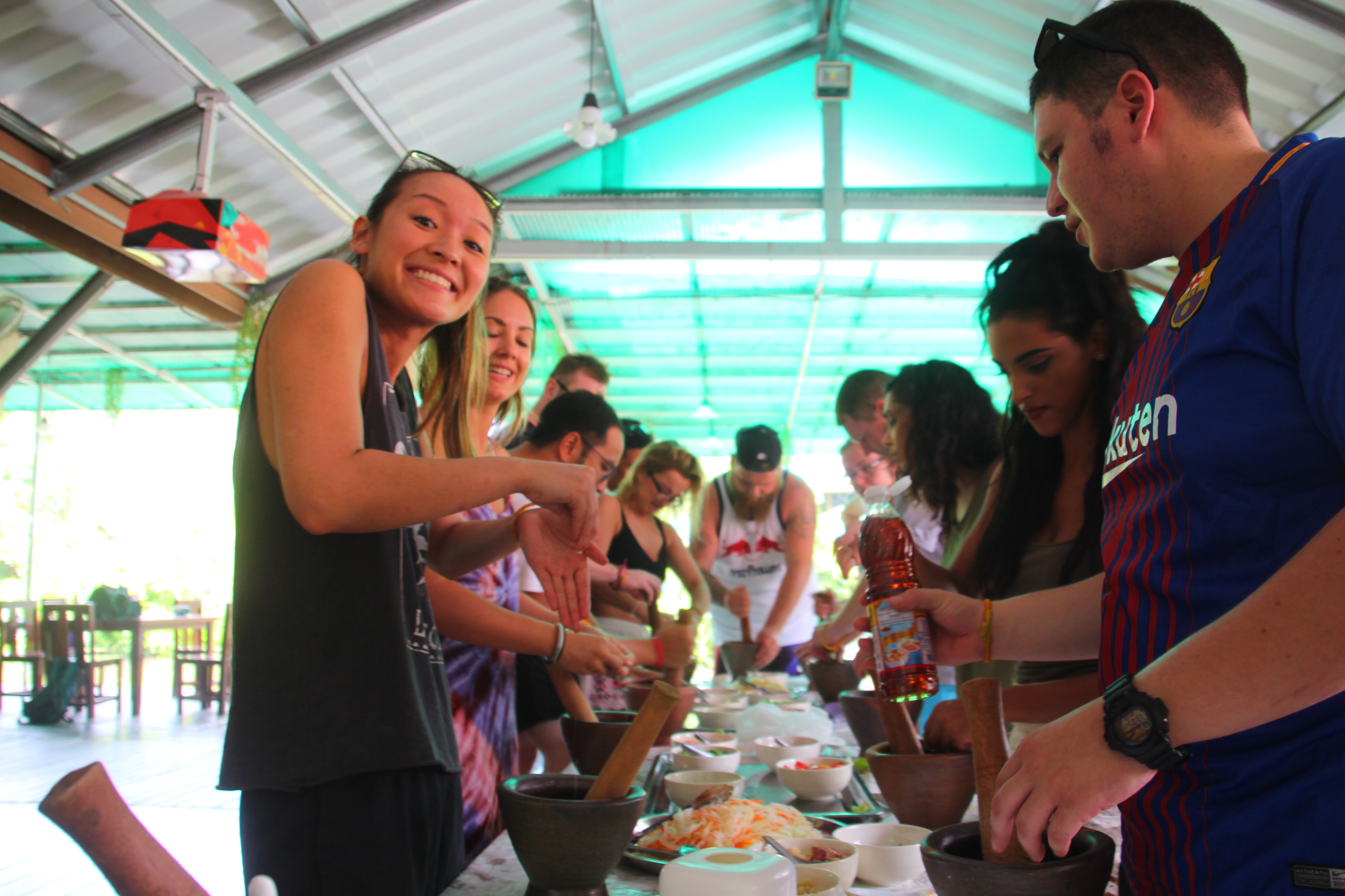 Smiling travelers learn to cook traditional Thai dishes using mortars and pestles in an open-air kitchen.