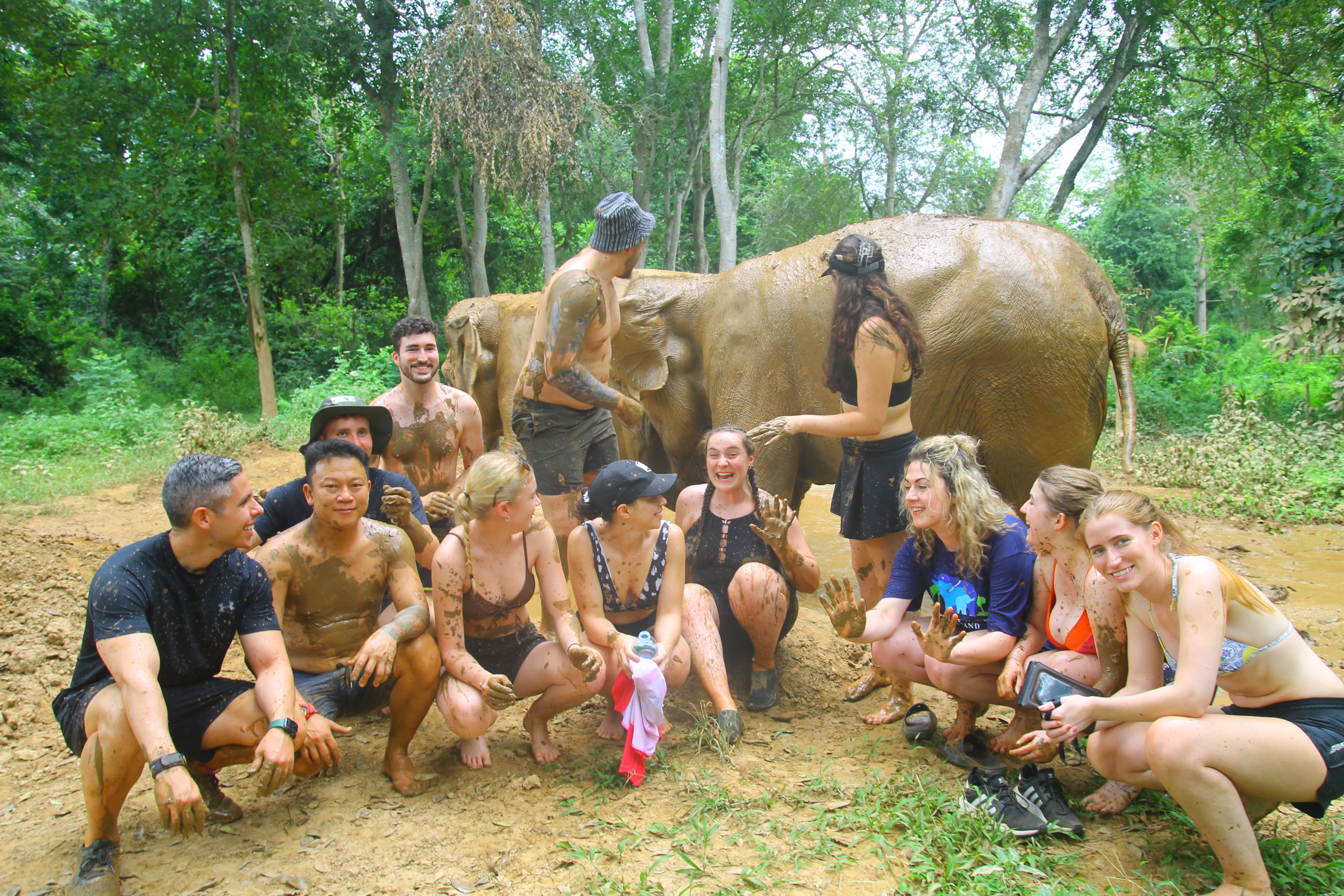 Happy tourists covered in mud with elephants in a lush Thai jungle, enjoying an adventurous experience.