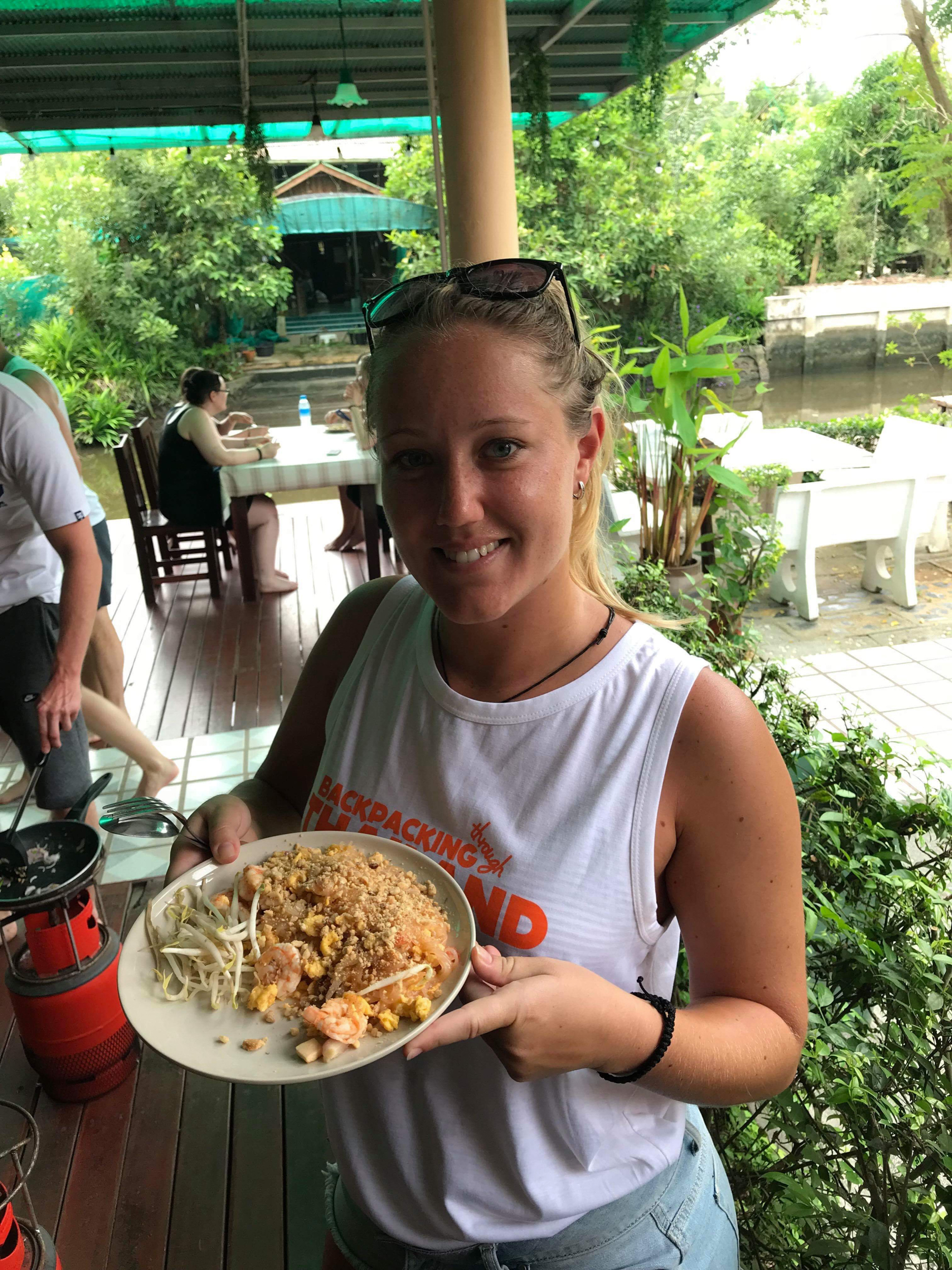 Happy backpacker holds a plate of Pad Thai at an outdoor cooking class in Thailand.