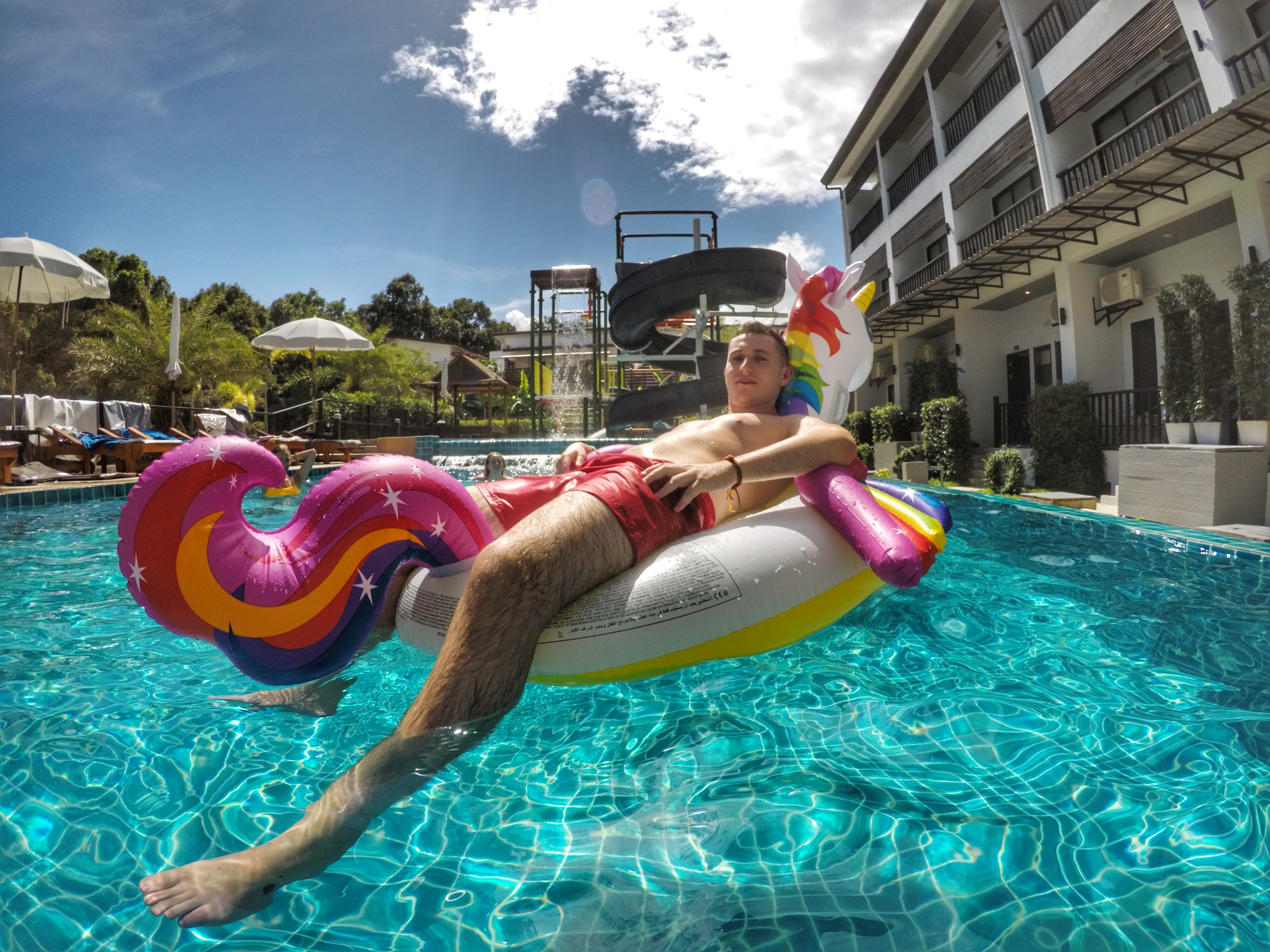 A man floats on a rainbow unicorn in a resort swimming pool under a blue sky.