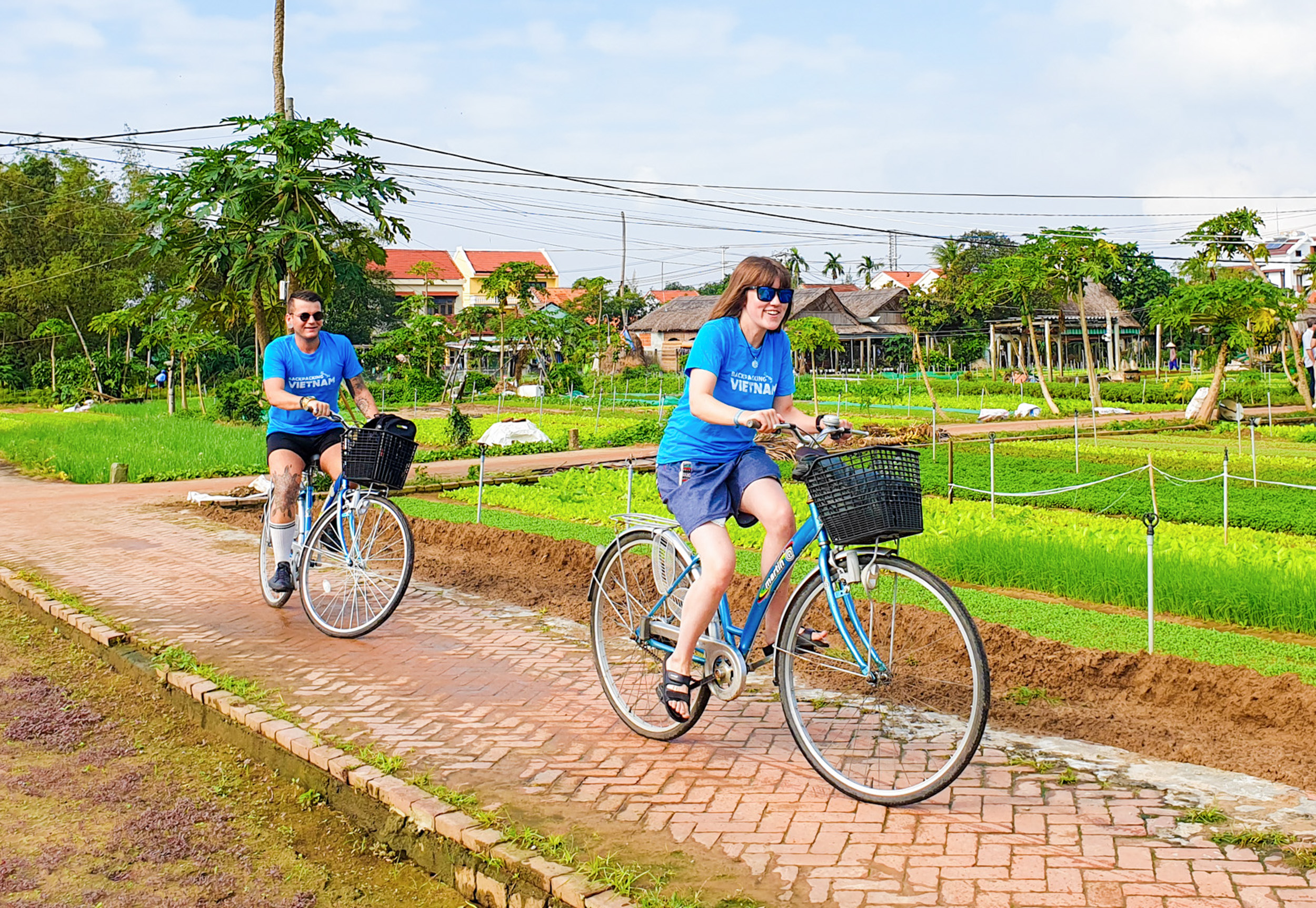 Two tourists happily cycle through lush green vegetable fields and traditional houses in Vietnam.