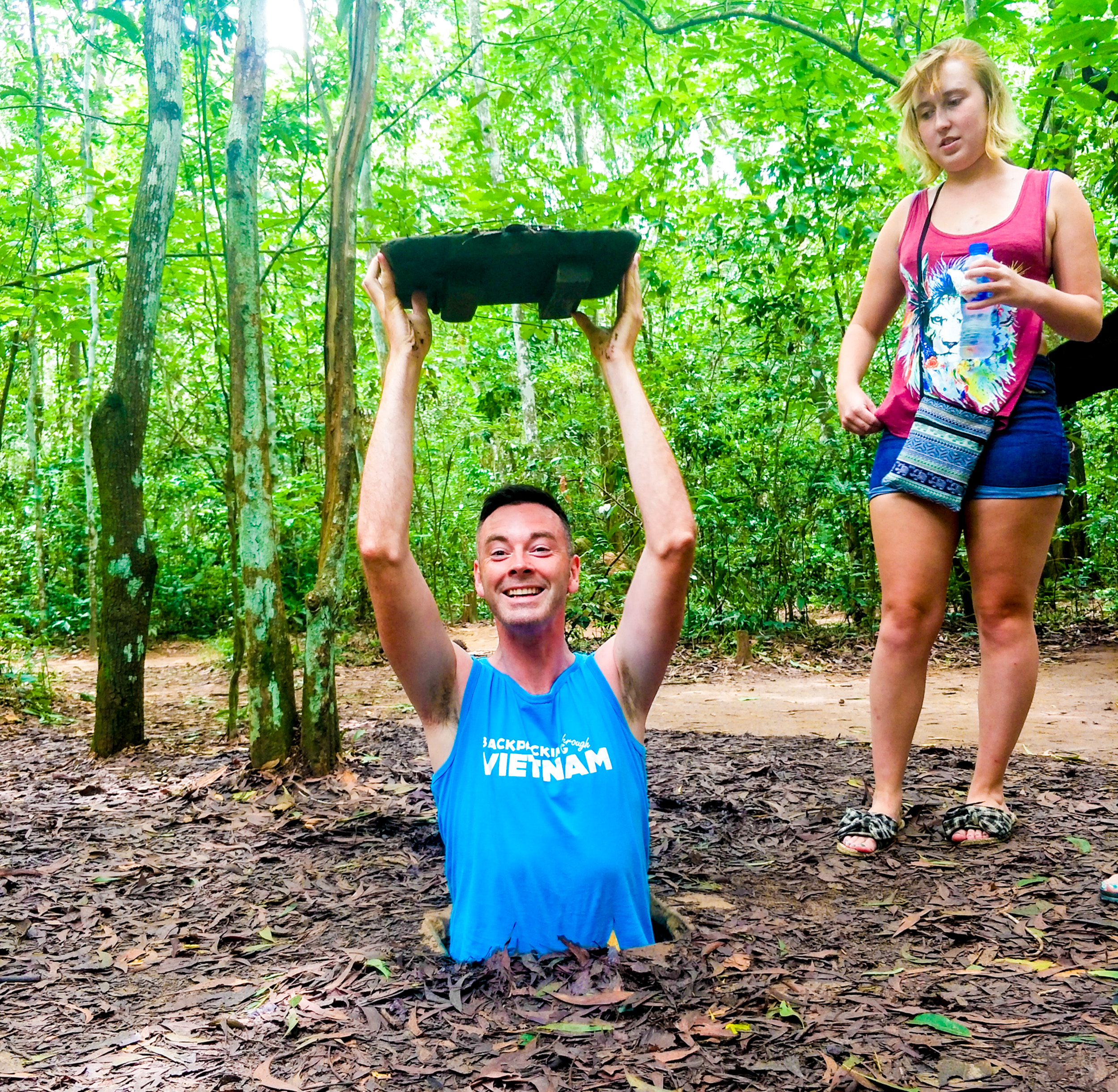 A smiling man emerges from a muddy tunnel, holding a lid, as a woman watches in Vietnam.