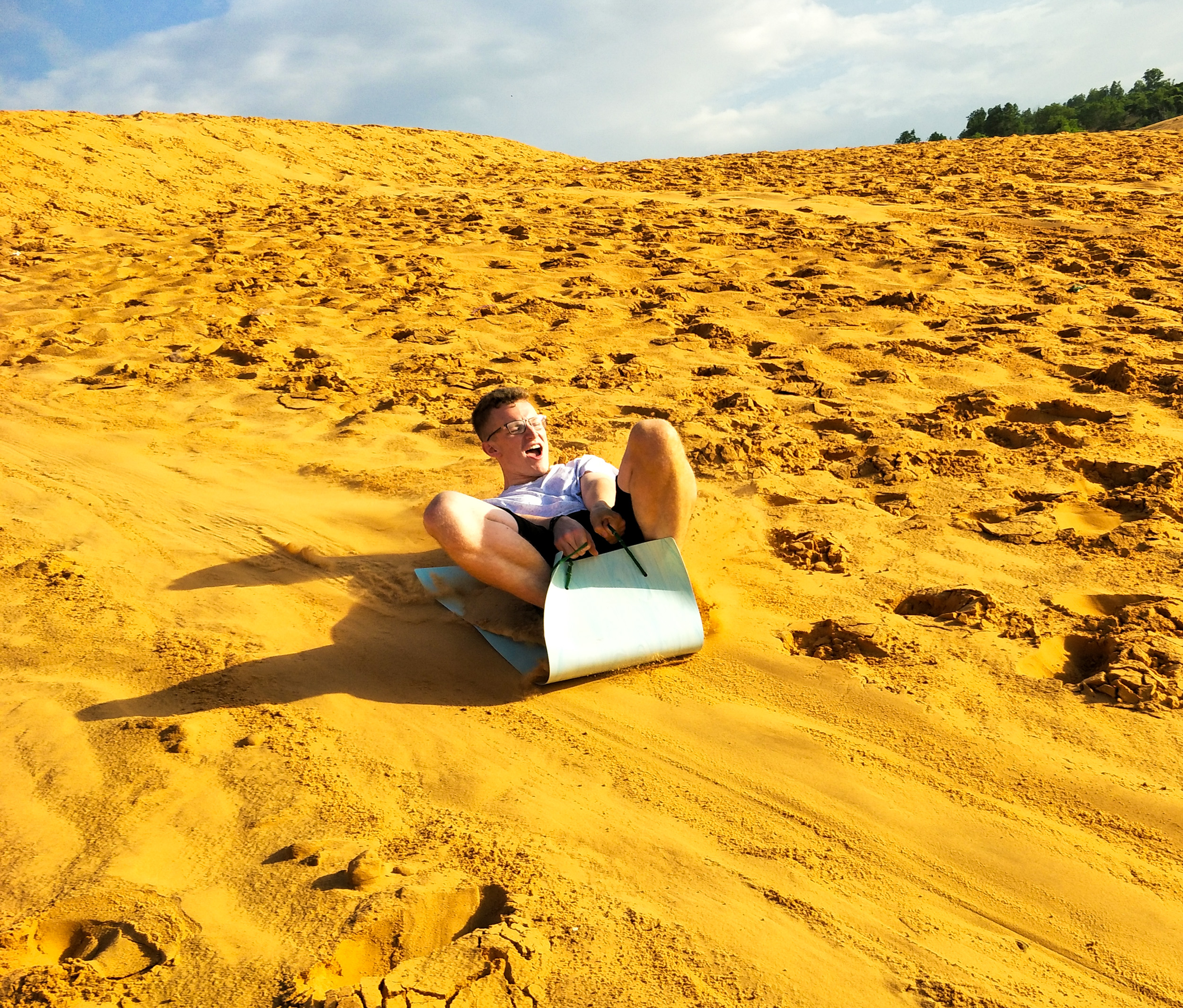 A young man smiles while sandboarding down a golden dune in Mui Ne, Vietnam.