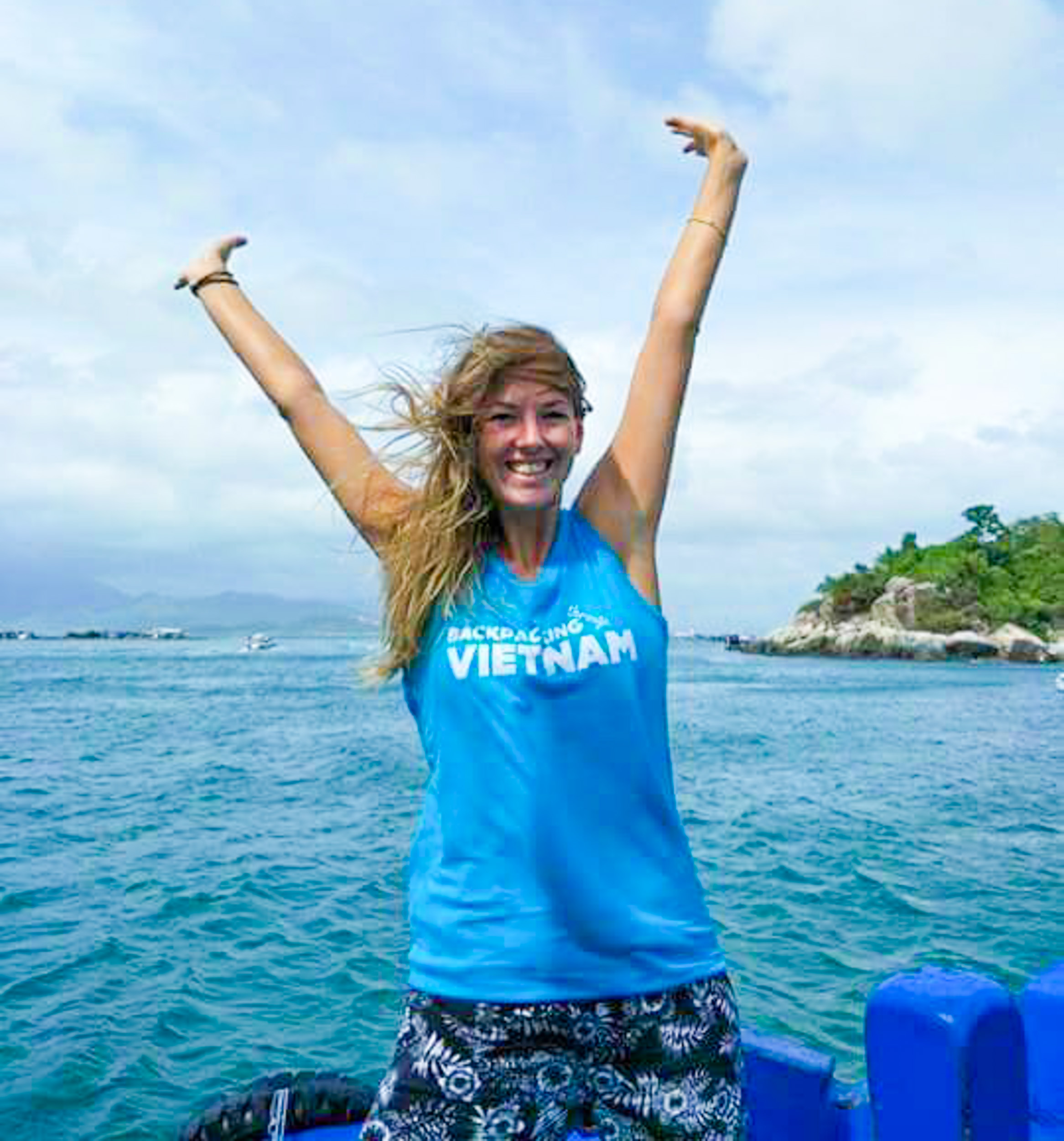Smiling woman in a 'Backpacking Vietnam' shirt with arms raised on a boat.